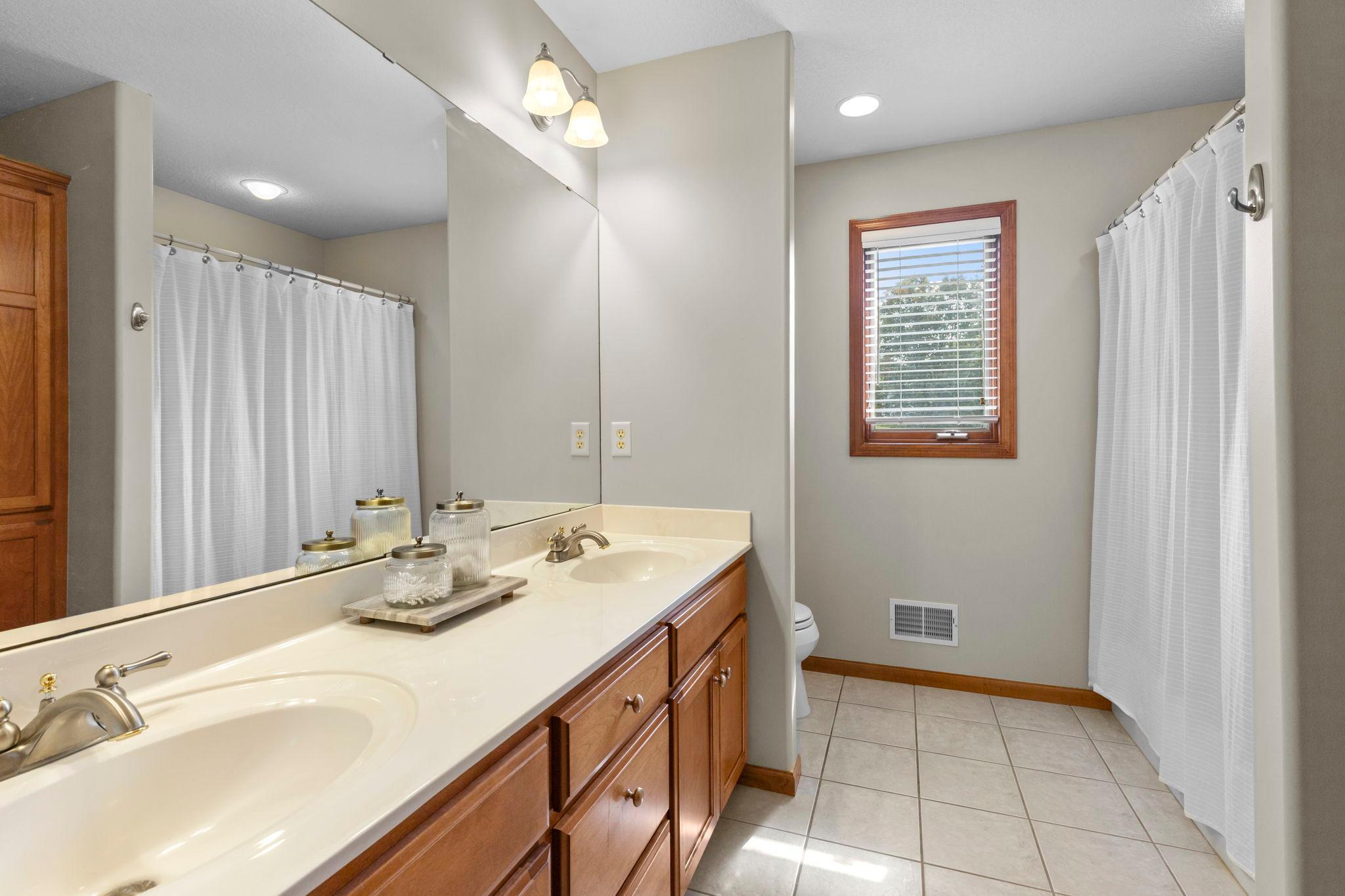Bathroom with a long double-sink wood vanity, large mirror, tiled floor, and window.