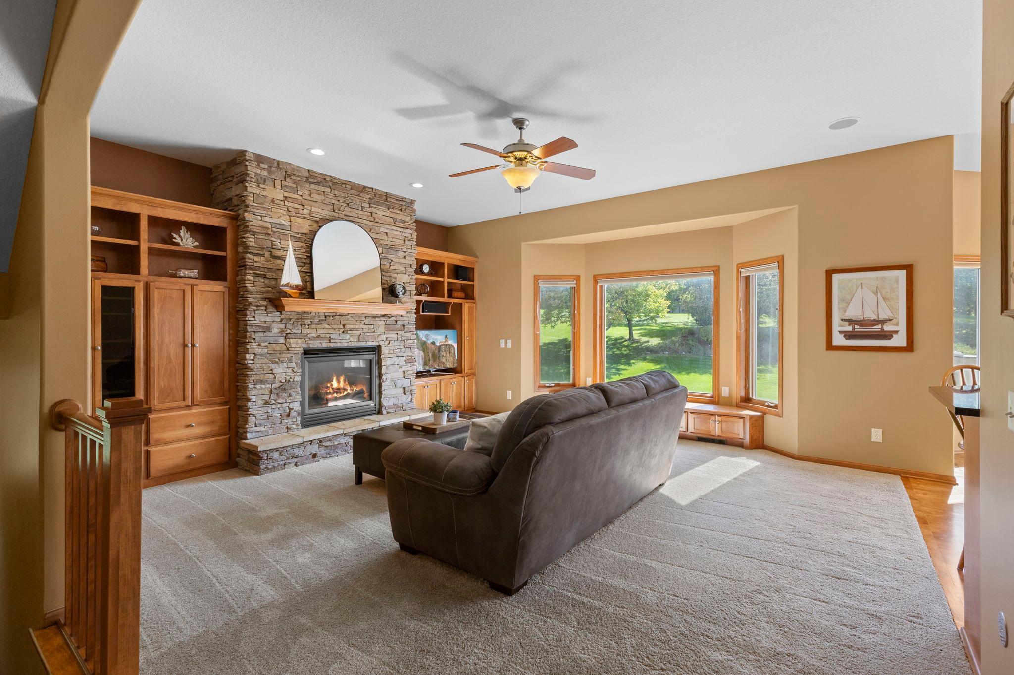 Living room with a stone fireplace, built-in wood cabinetry, and large windows overlooking a lawn.