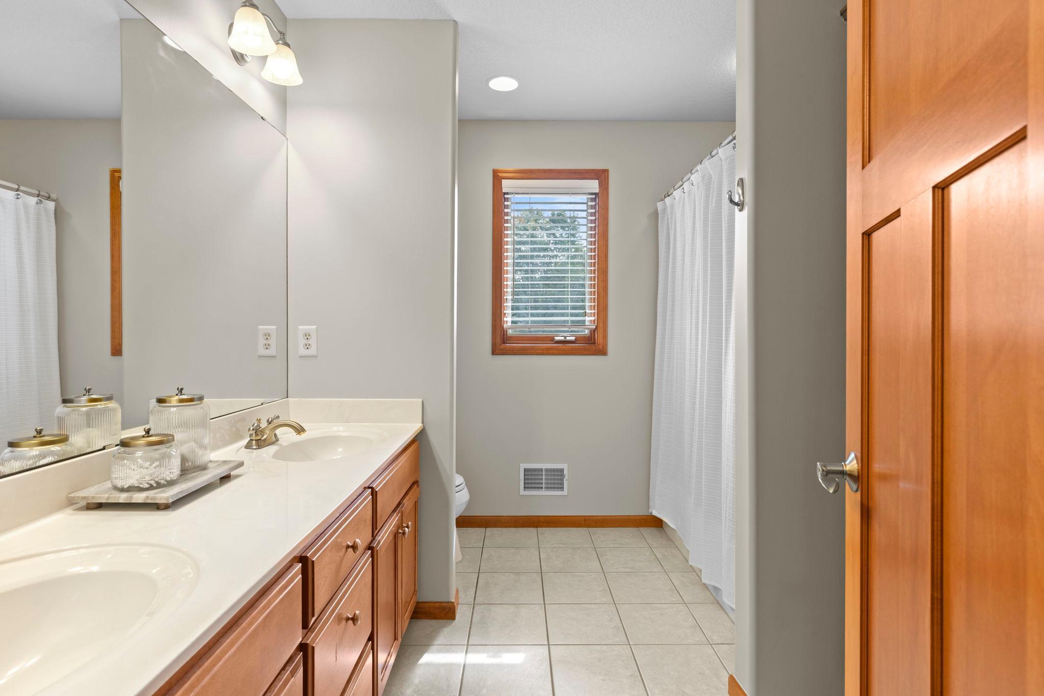 Bathroom with a long wood double vanity, tiled floor, and a window beside the curtained tub.