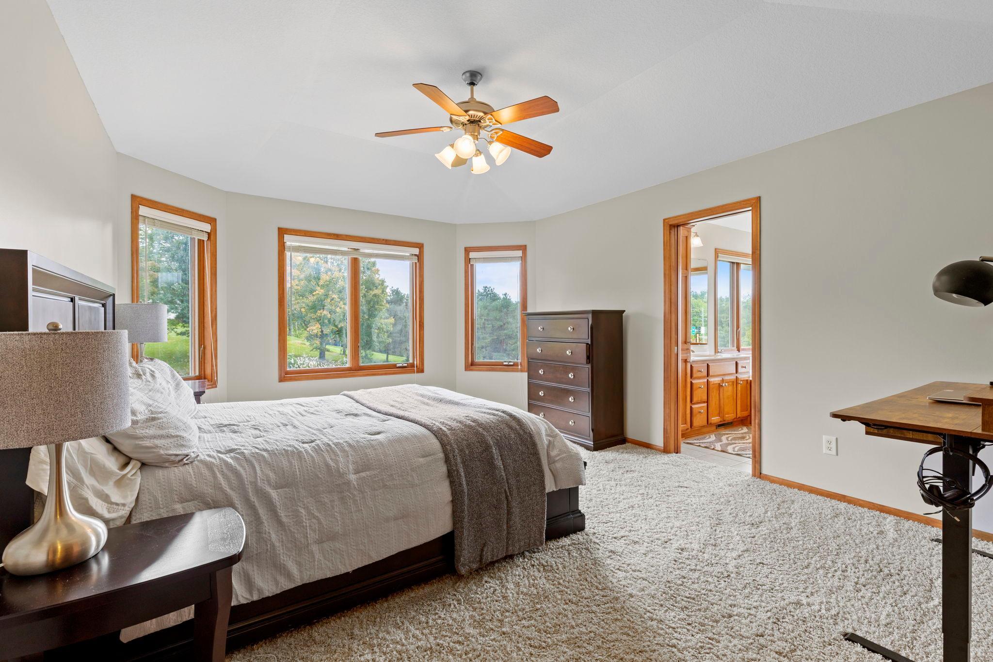 Bedroom with a large bed, wood-trimmed windows overlooking trees, and an adjoining bathroom.