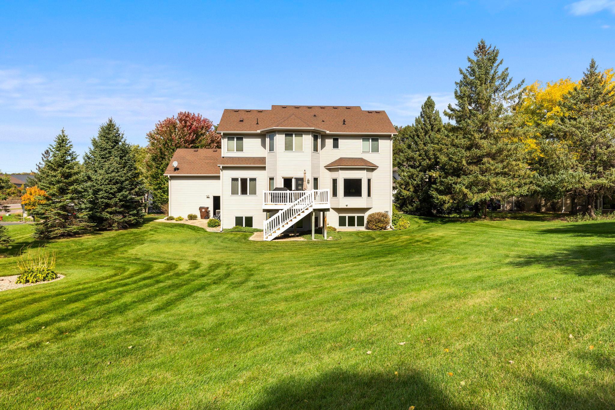 Rear of a two-story home with white deck stairs and a broad lawn framed by mature trees.