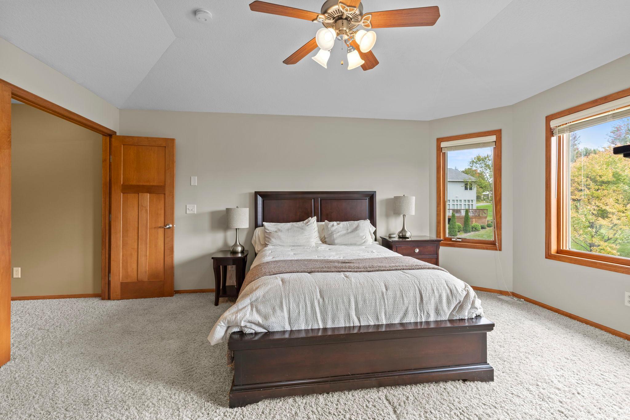 Bedroom with a dark wood bed, white bedding, and two windows overlooking trees and a neighboring house.