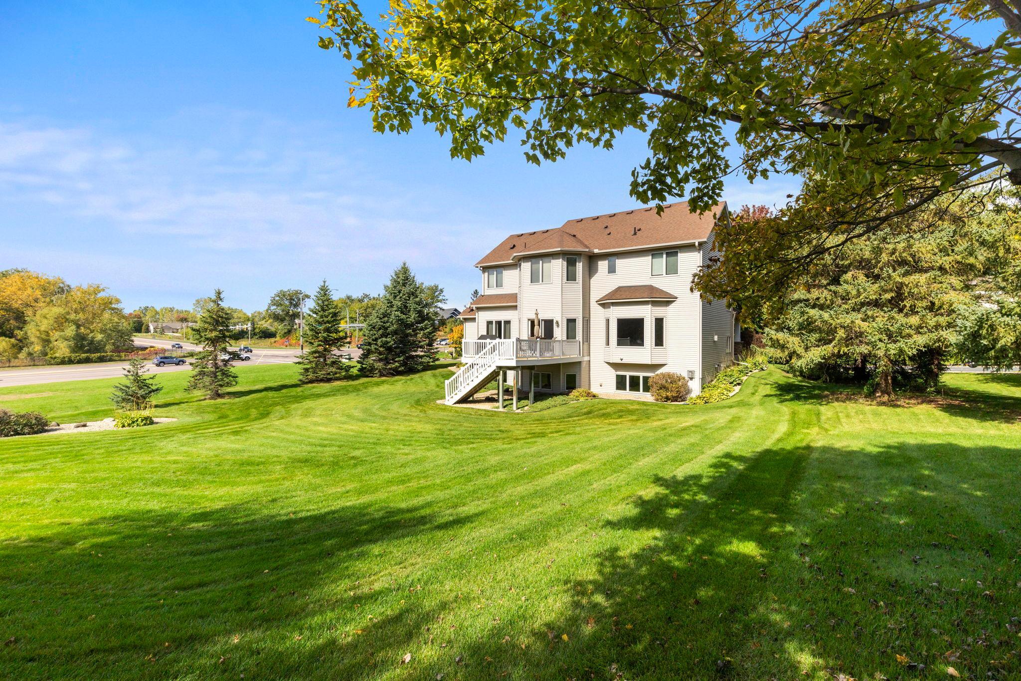 Two-story home with a raised deck and white stairs beyond a broad lawn and mature trees.