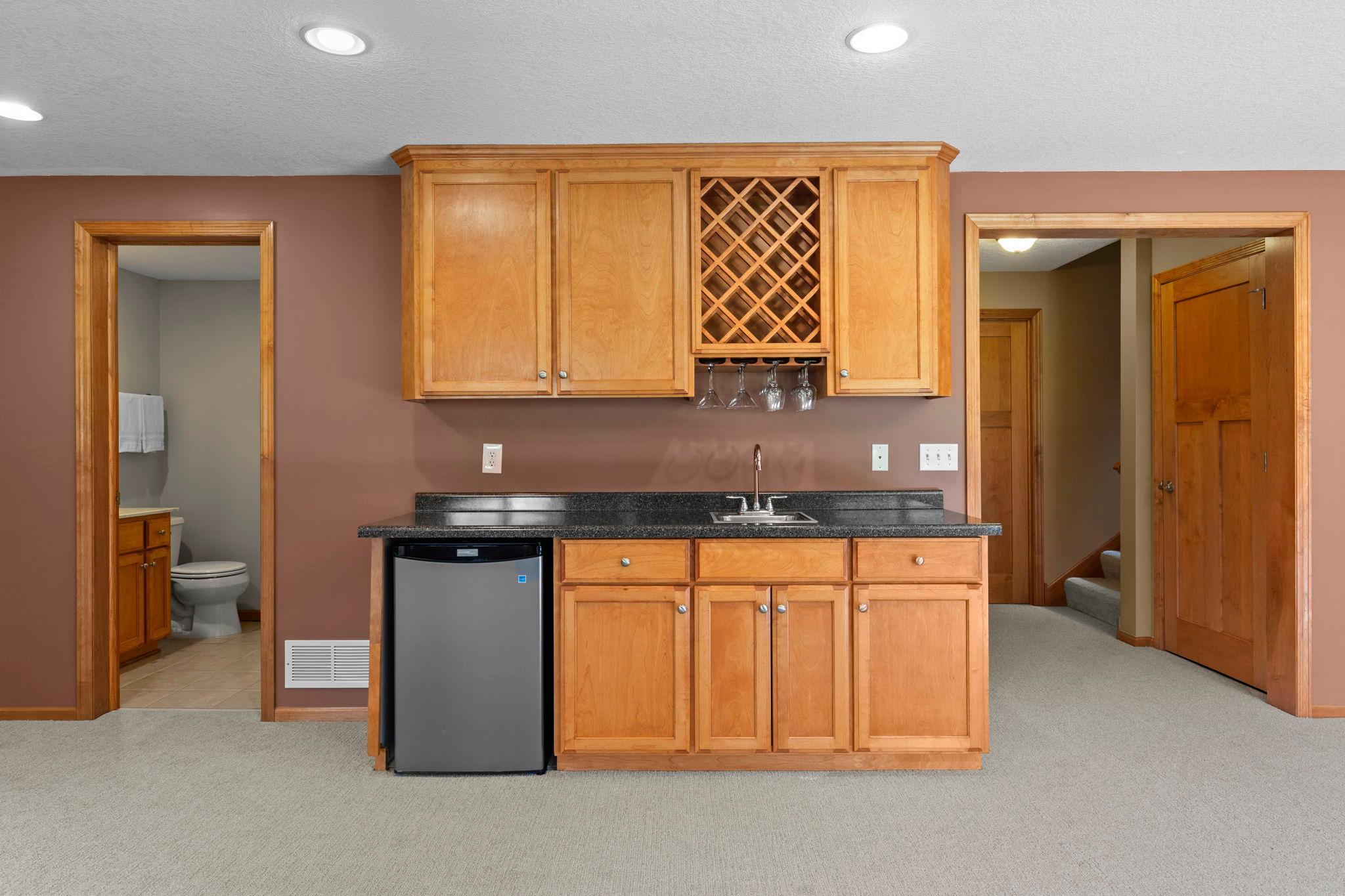 Wet bar with wood cabinetry, lattice wine rack, black countertop, sink, and compact refrigerator.