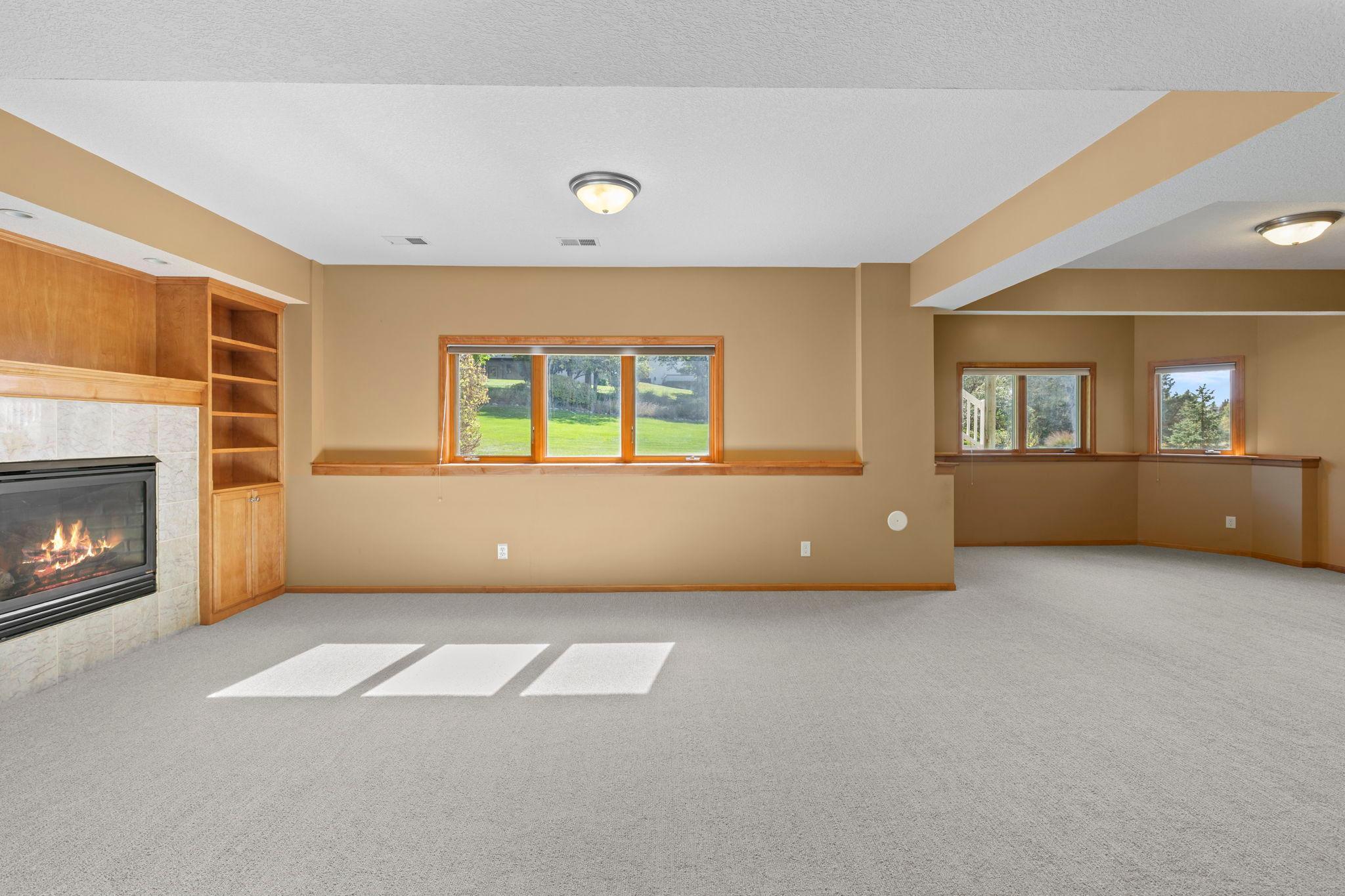 Empty living room with a tiled fireplace, built-in wood shelving, and several sunlit windows.