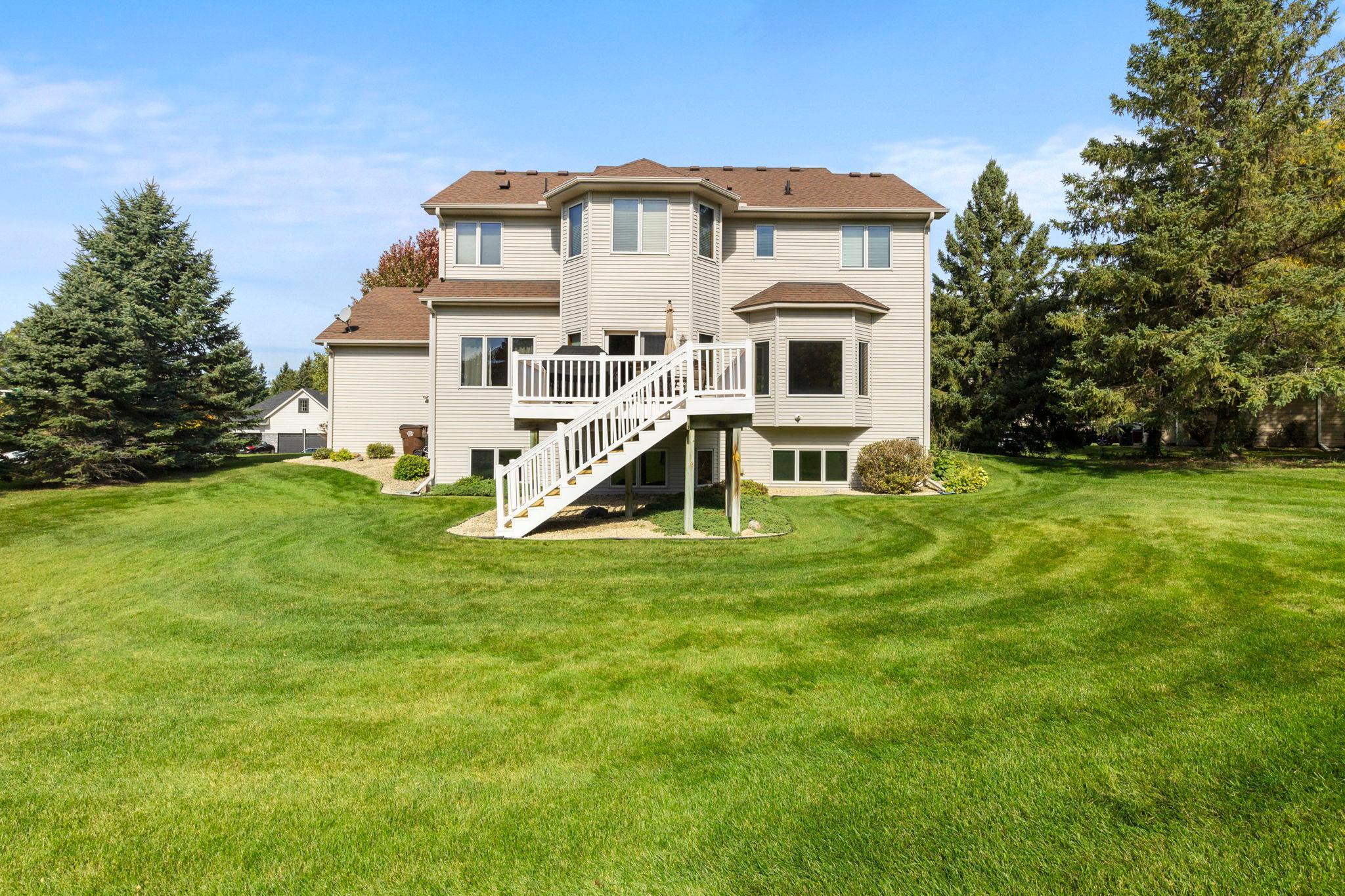 Rear of a two-story beige house with a white deck and stairs overlooking a broad lawn.