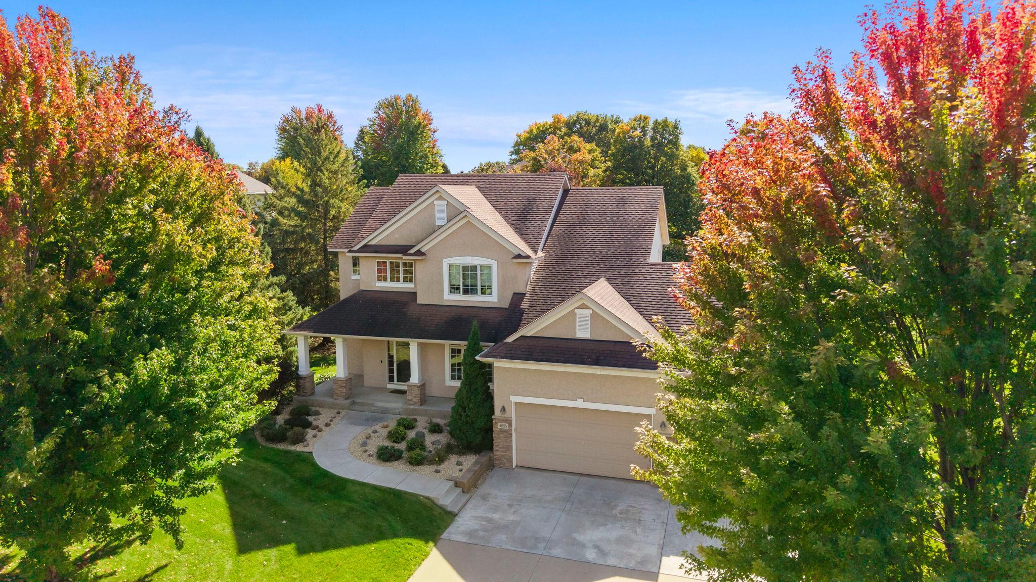 Two-story beige home with an attached garage and covered porch, framed by colorful autumn trees.