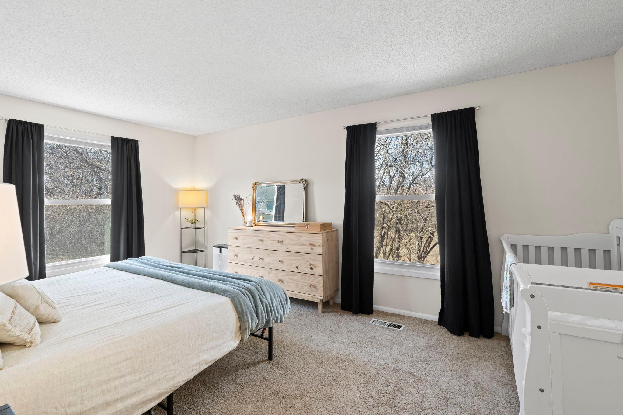 Bedroom with a white bed, pale blue throw, light wood dresser, and white crib beside two windows.
