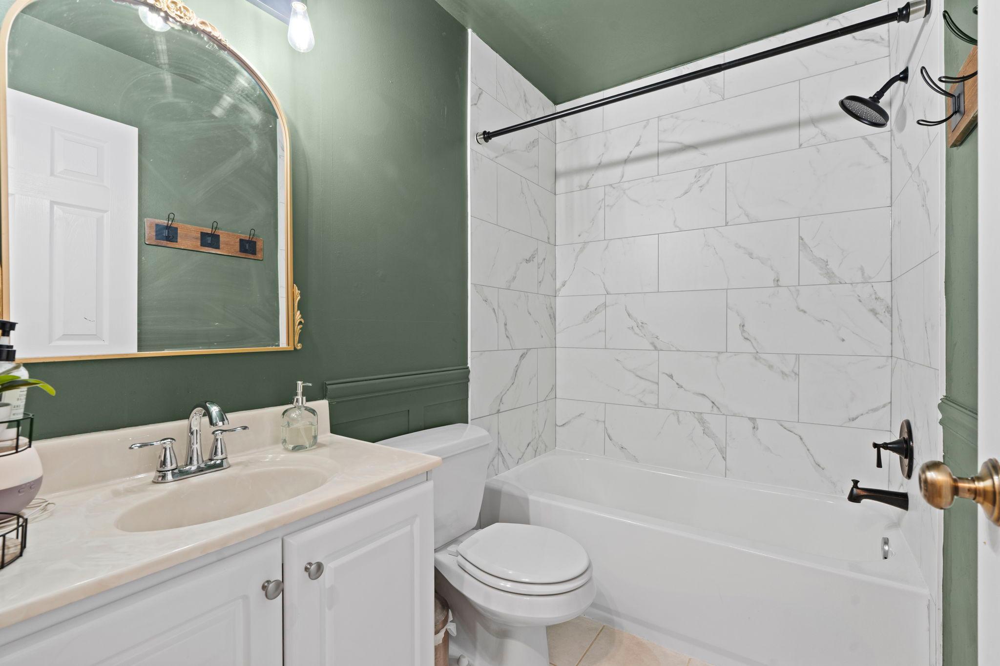 Bathroom with a marble-look tiled tub surround, dark green walls, and an arched gold-framed mirror.