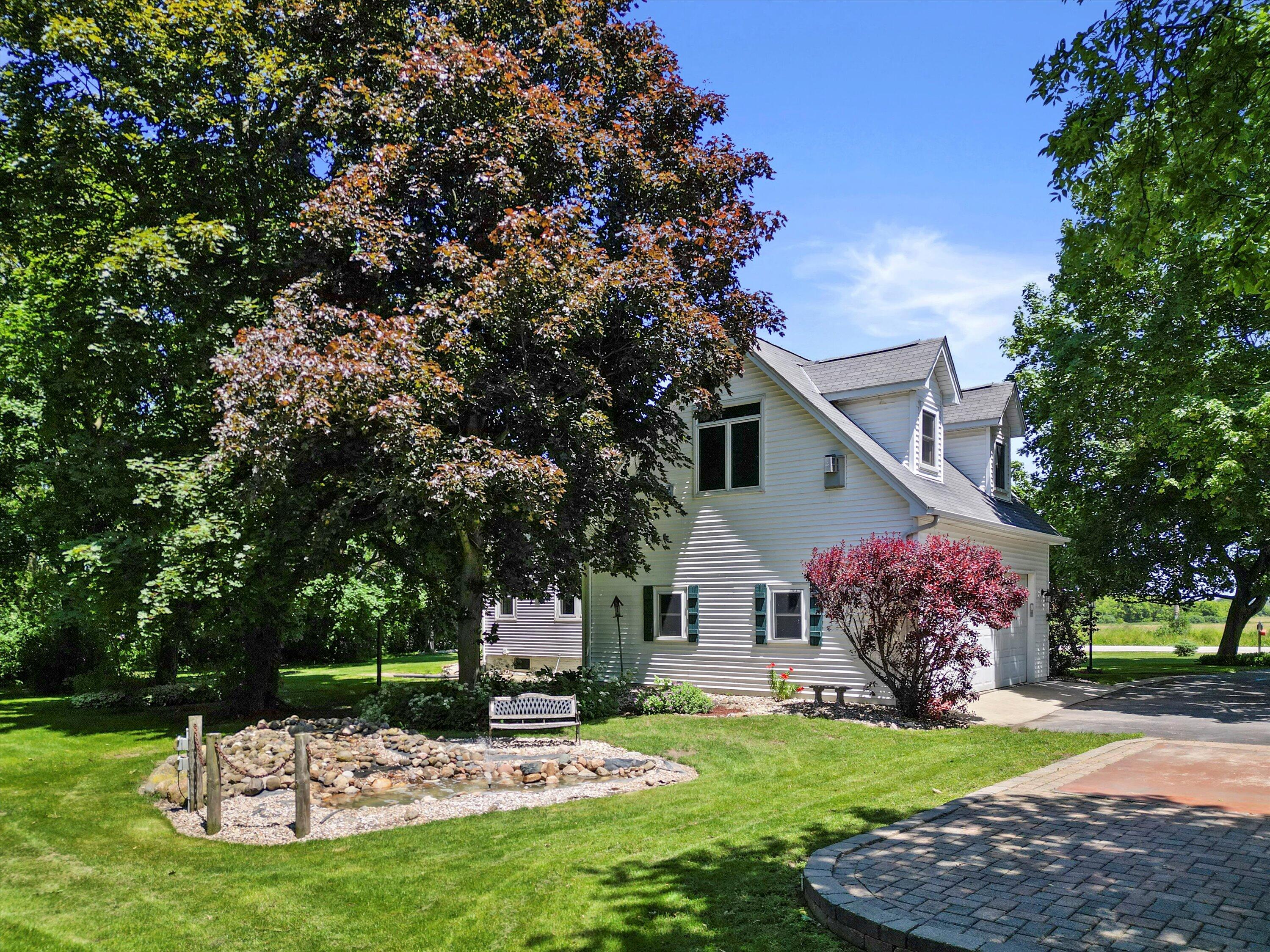 White two-story house among mature trees, with a rock-lined water feature and curved paver patio.
