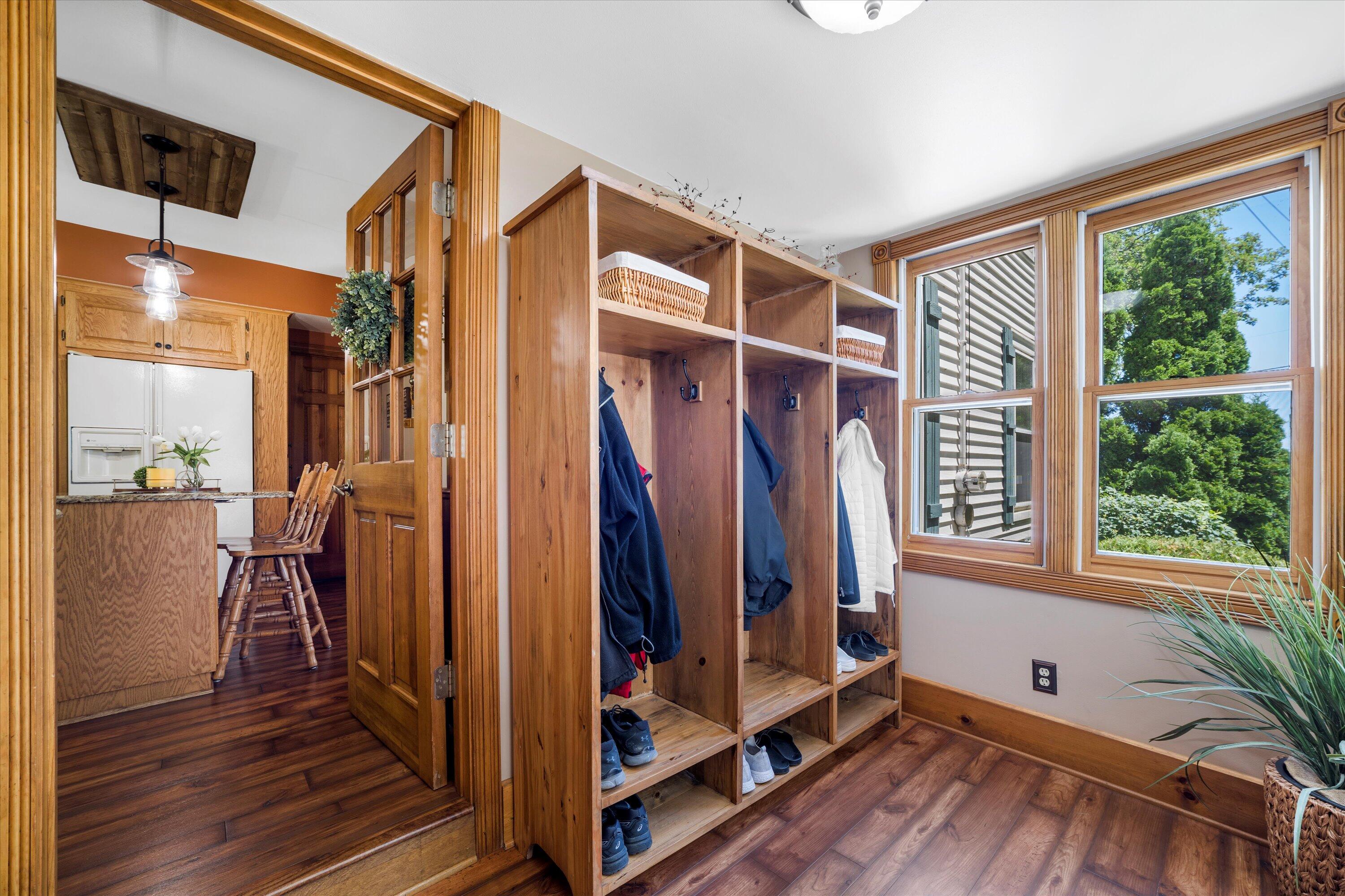 Mudroom with built-in wooden cubbies, coats and shoes, beside large windows and an open kitchen doorway.