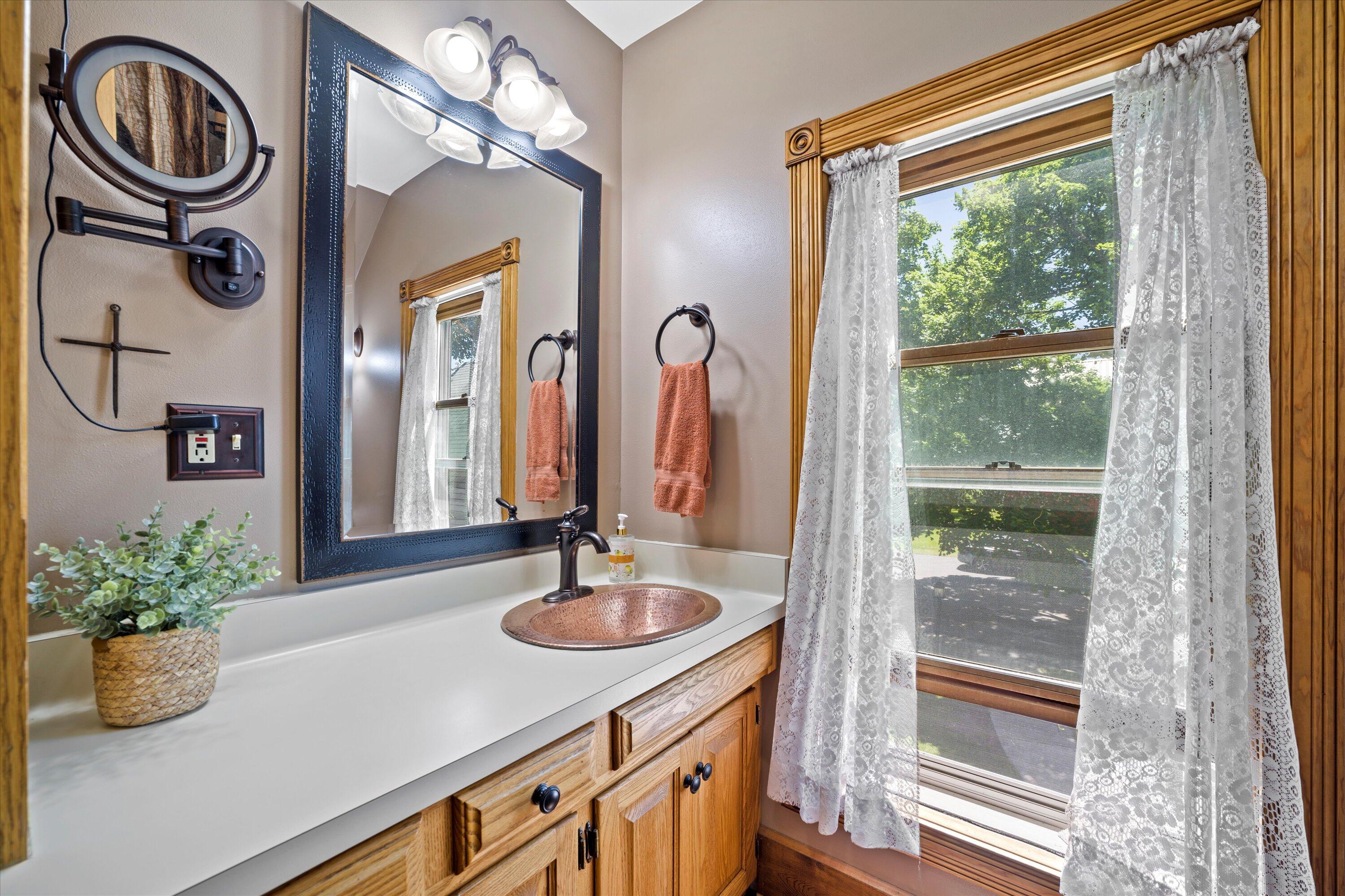 Bathroom vanity with a copper vessel sink, wood cabinetry, and a lace-curtained window.