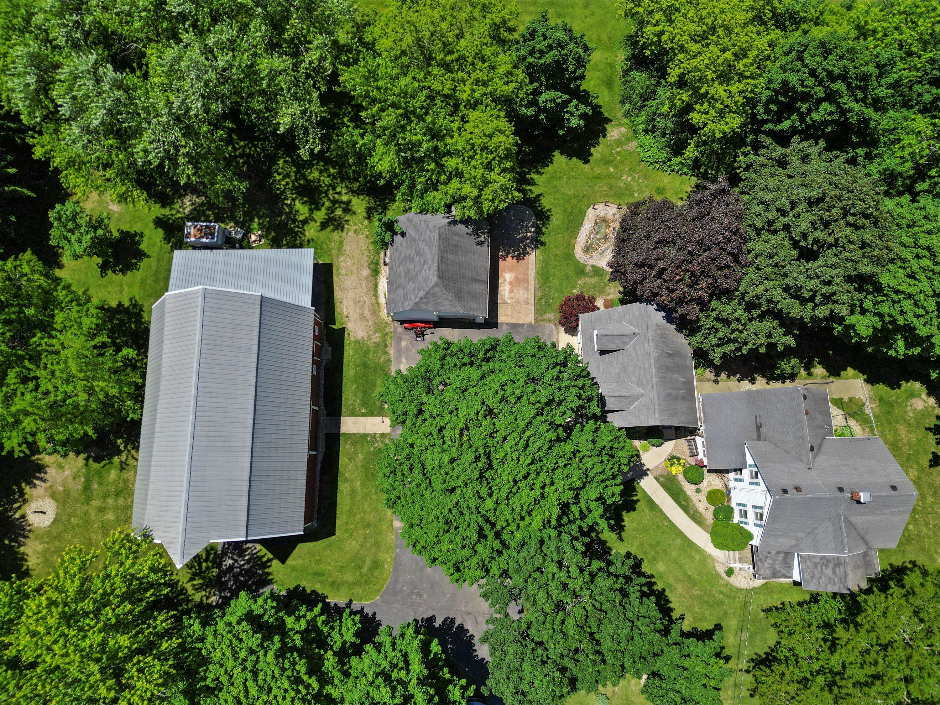 Aerial view of a house and detached outbuilding among mature trees, with a paved driveway and landscaped yard.