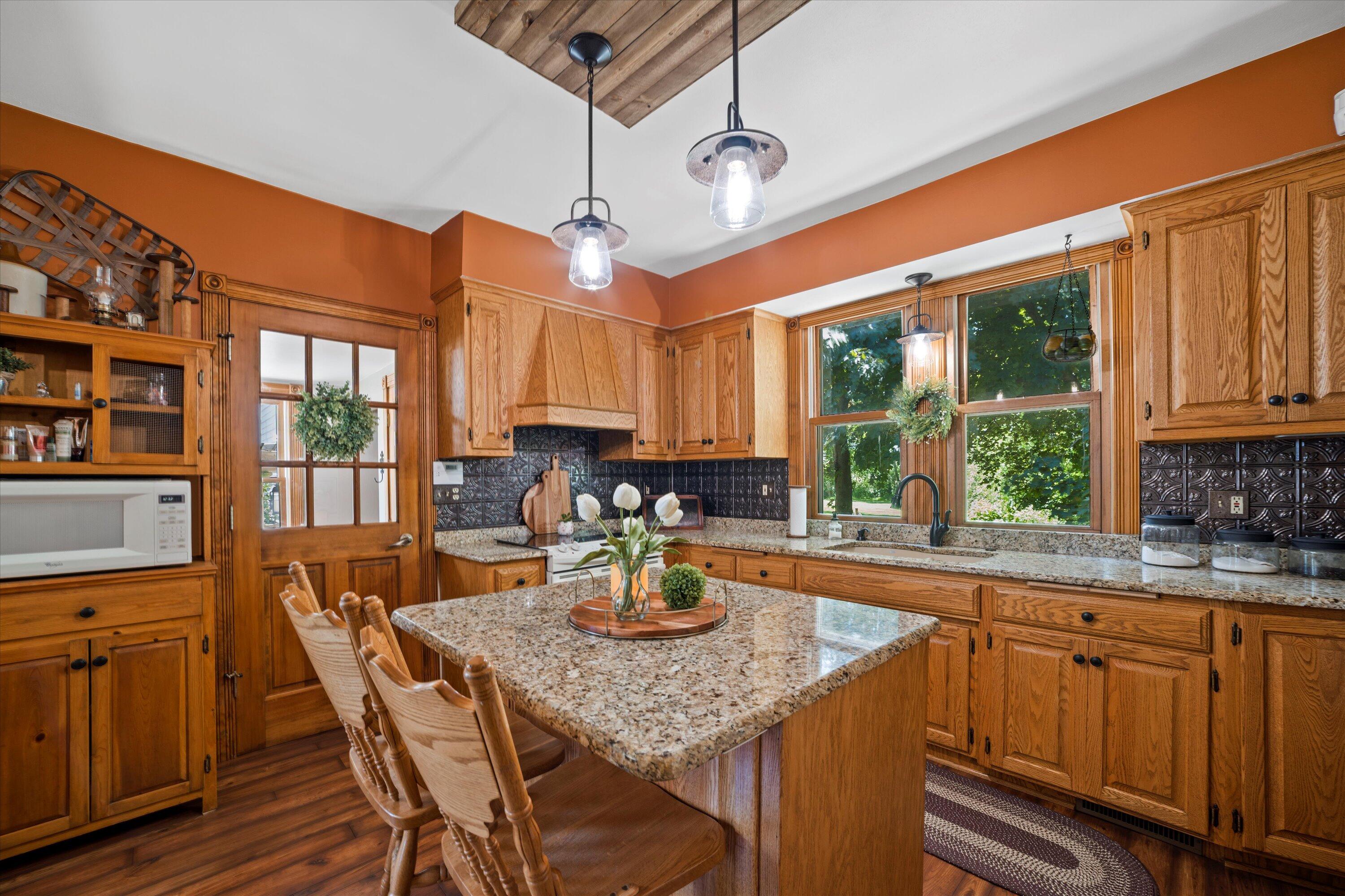 Kitchen with warm wood cabinets, granite island, dark tile backsplash, and large windows overlooking greenery.