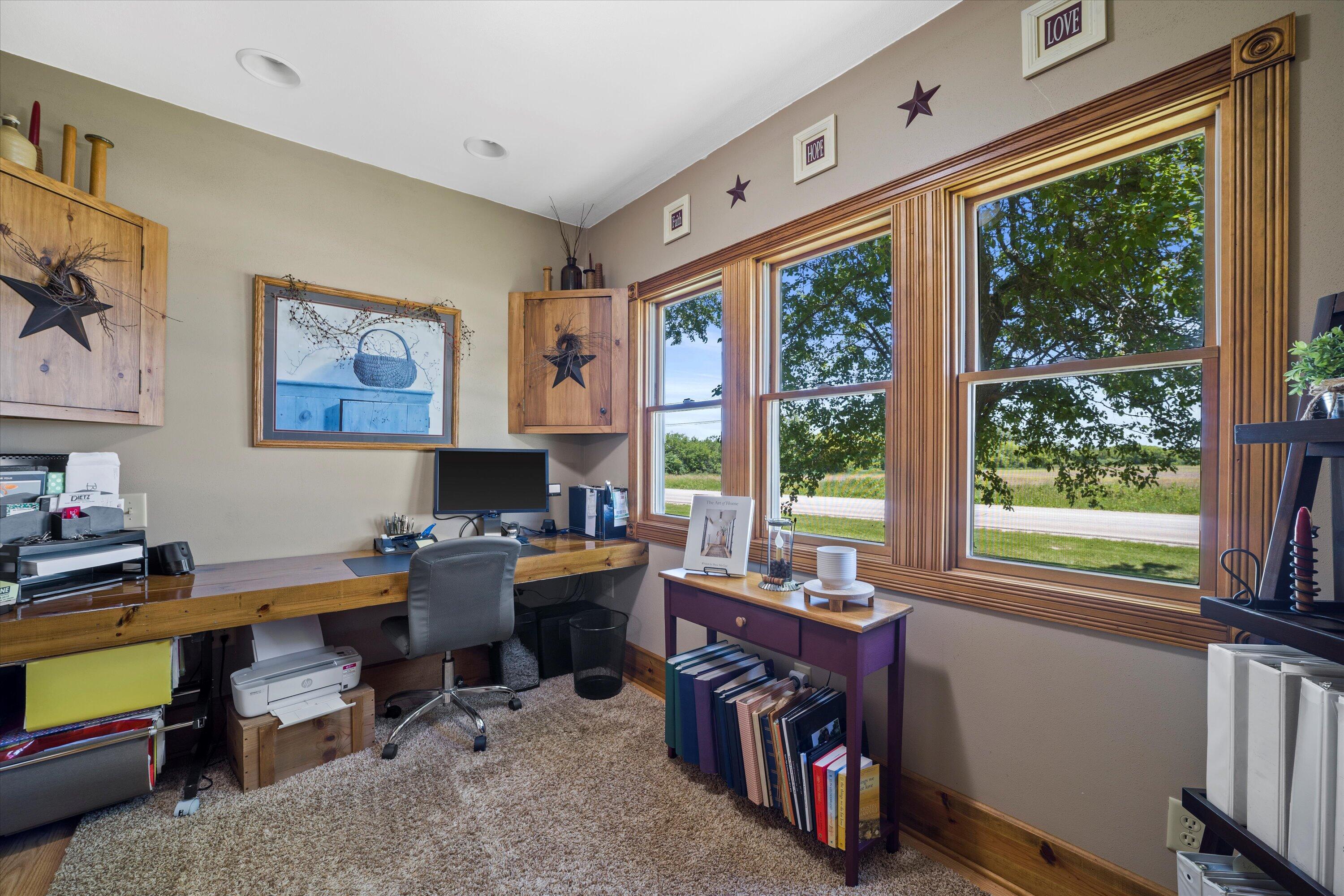 Home office with a long wood desk and large wood-trimmed windows overlooking a tree and road.