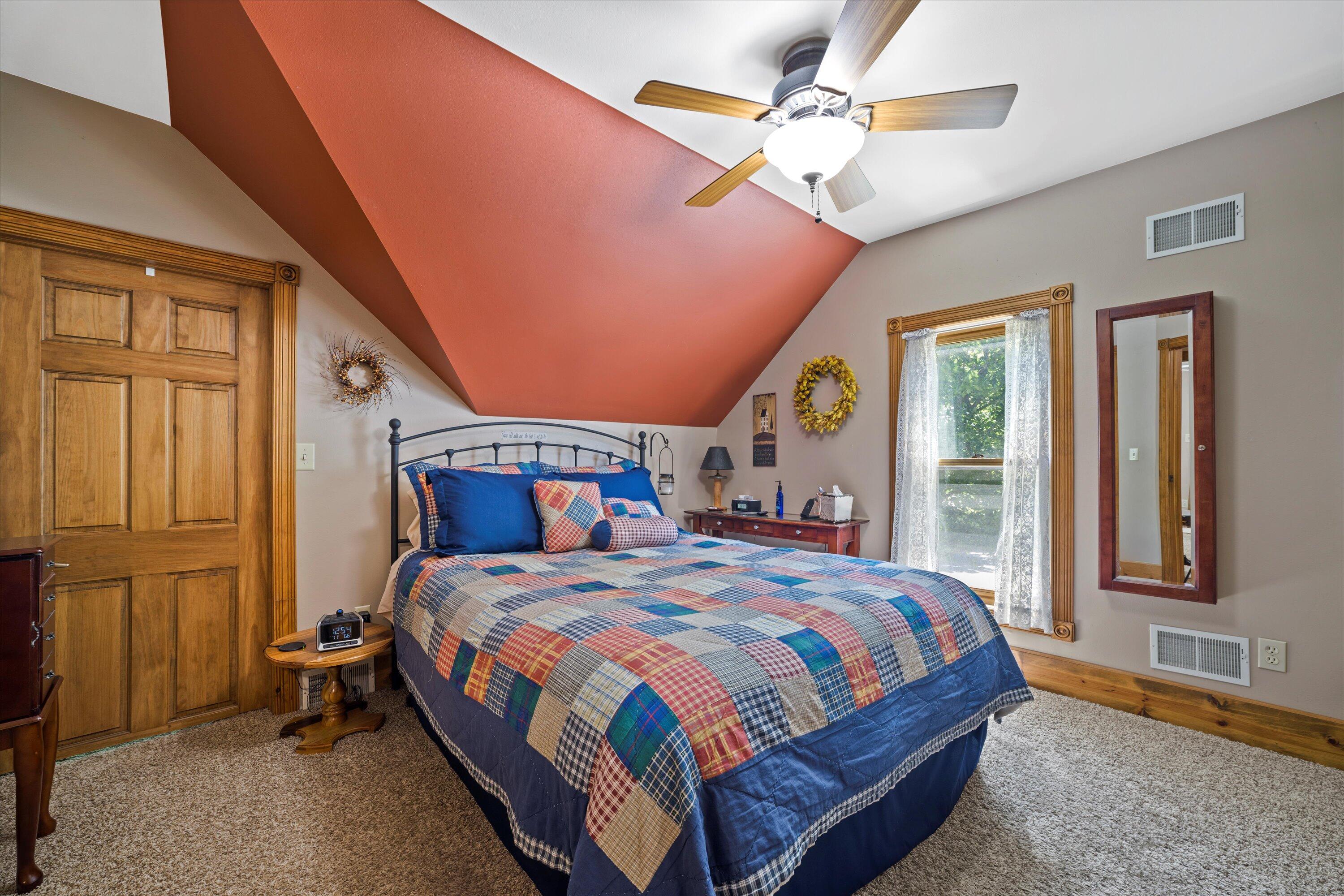 Furnished bedroom with a patchwork quilt, sloped rust-colored ceiling, ceiling fan, and wood-trimmed doors.