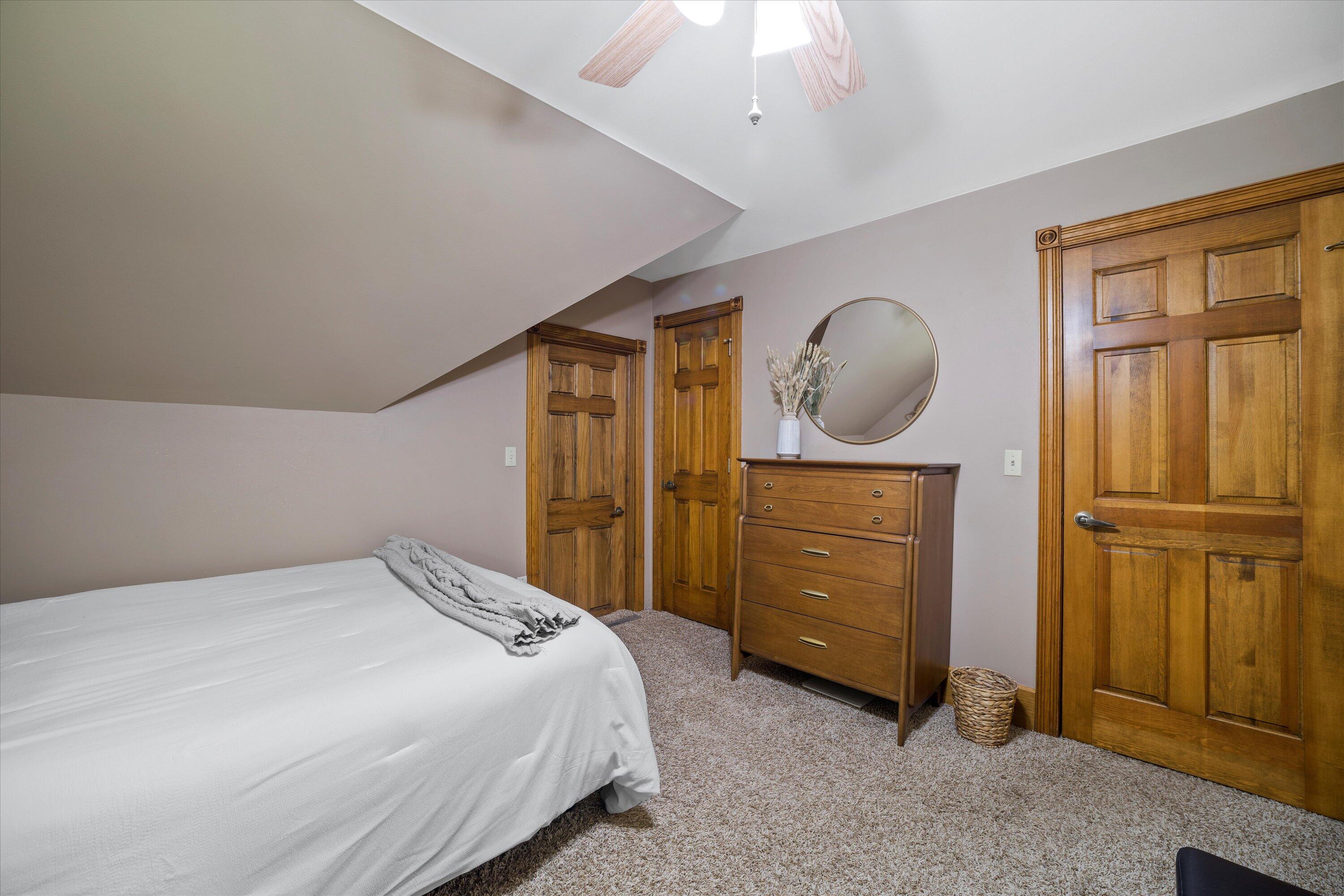 Bedroom with a sloped ceiling, white-covered bed, wooden dresser, and paneled wood doors.