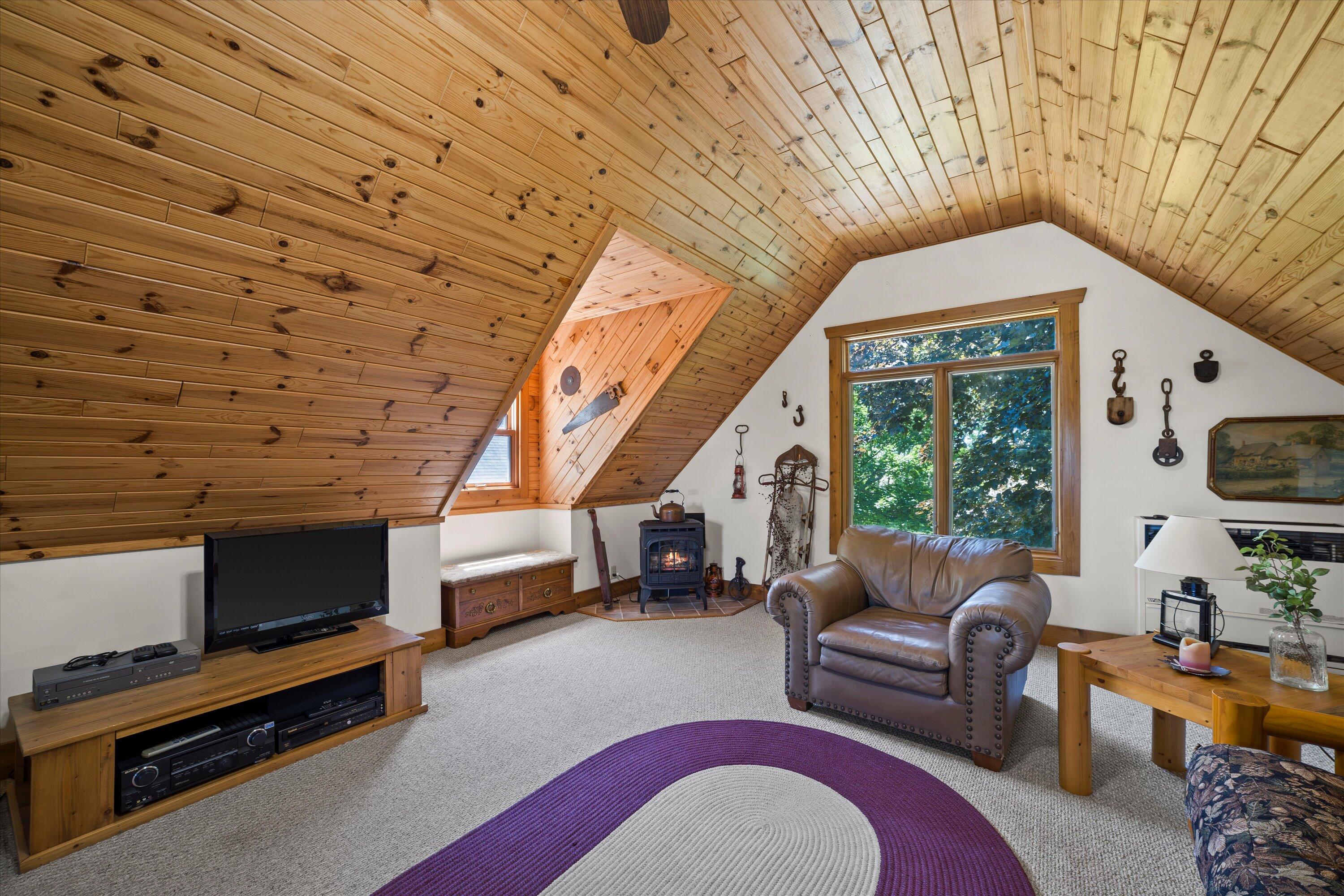 Attic living room with a knotty-pine ceiling, wood stove, and large window overlooking trees.
