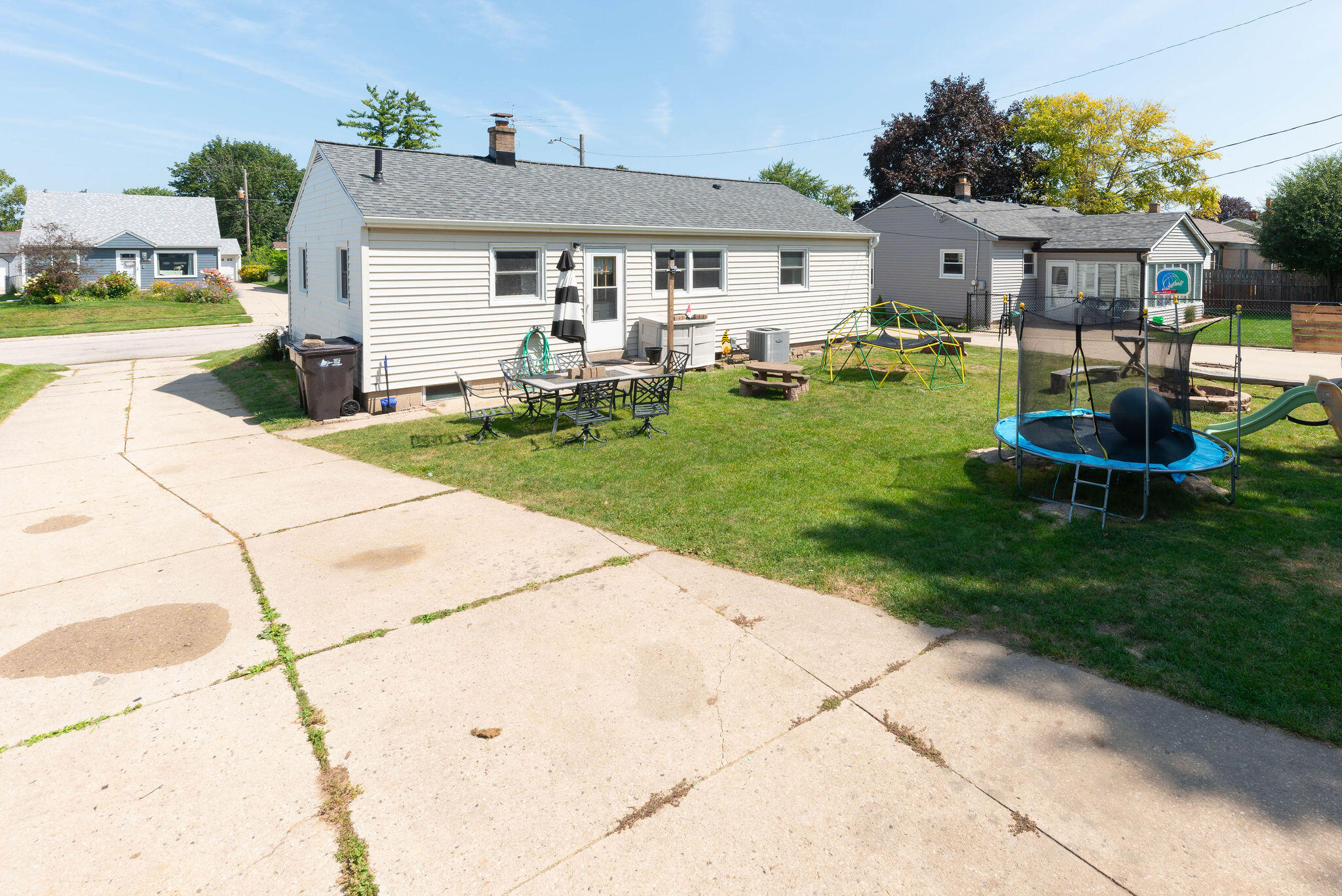 Rear of a single-story white house with a grassy yard, patio furniture, climbing dome, and trampoline.