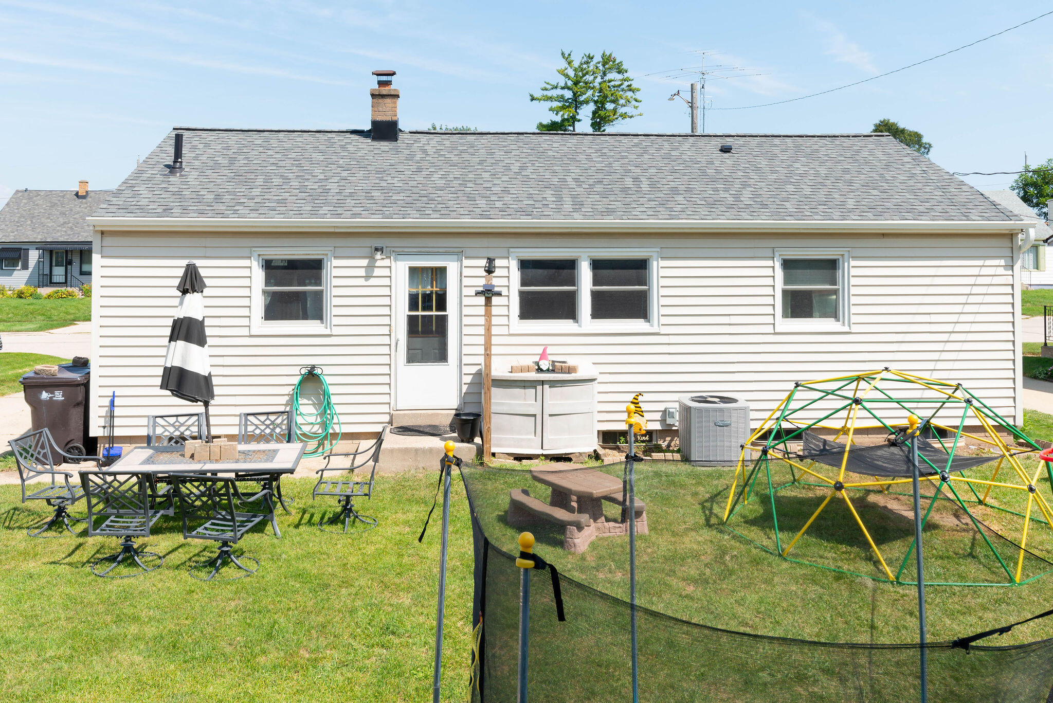White-sided home with a gray roof and backyard patio furniture, climbing frame, and trampoline.