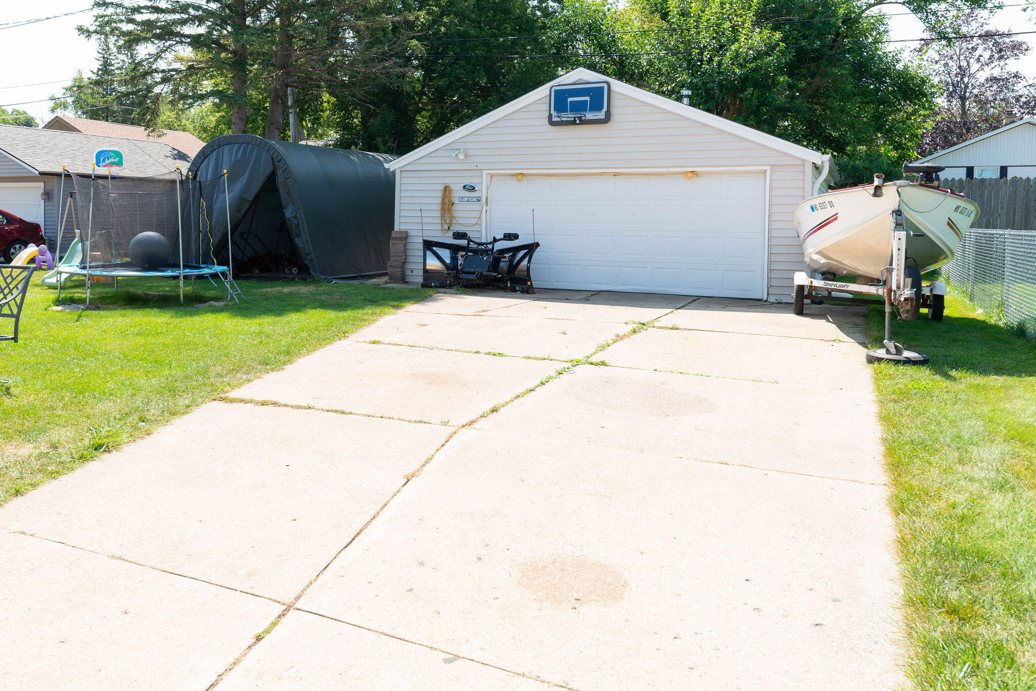 White detached garage with a basketball hoop, a wide driveway, trampoline, and boat on a trailer.
