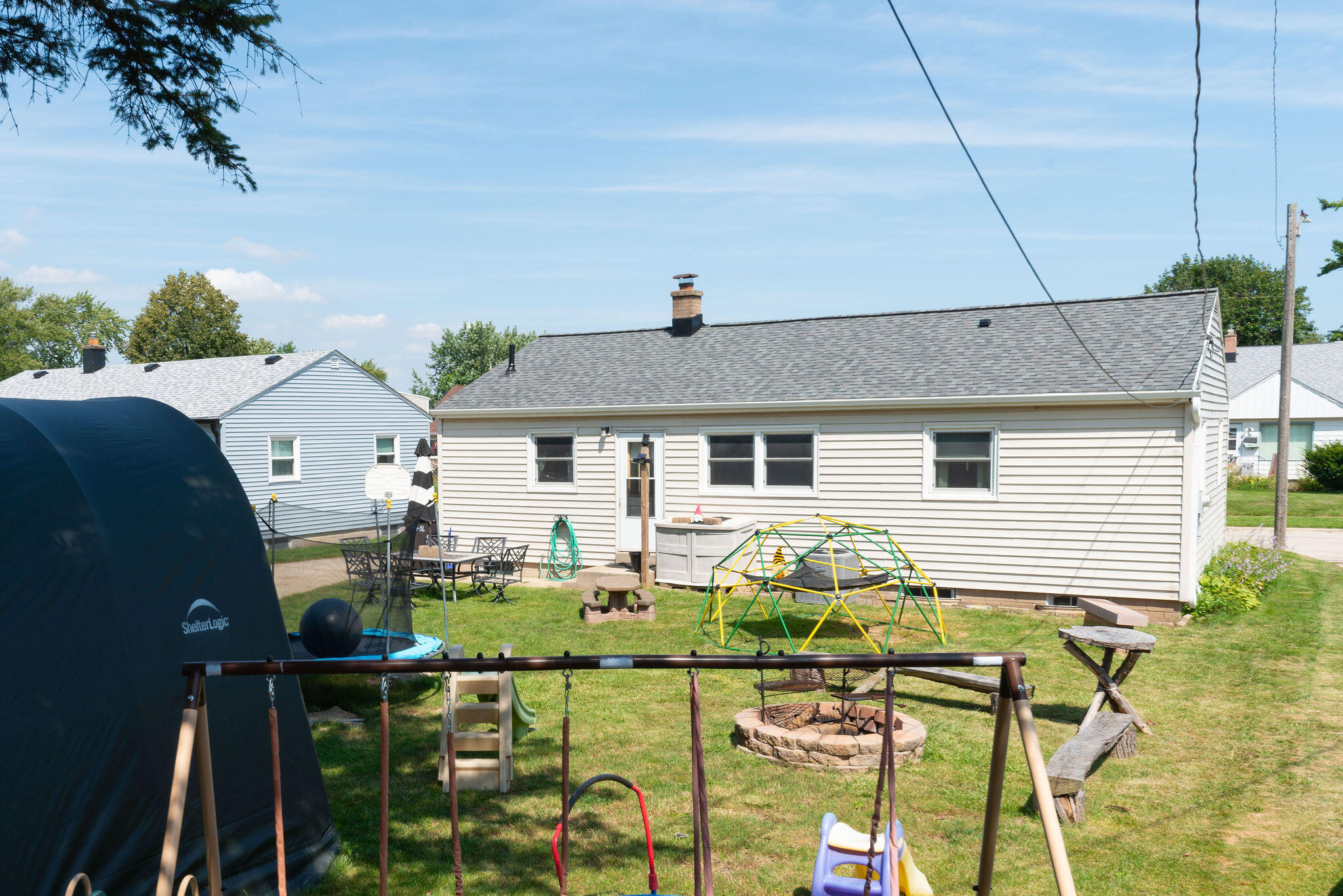 Rear of a white-sided house overlooking a backyard with play equipment, a fire pit, and a covered pool.