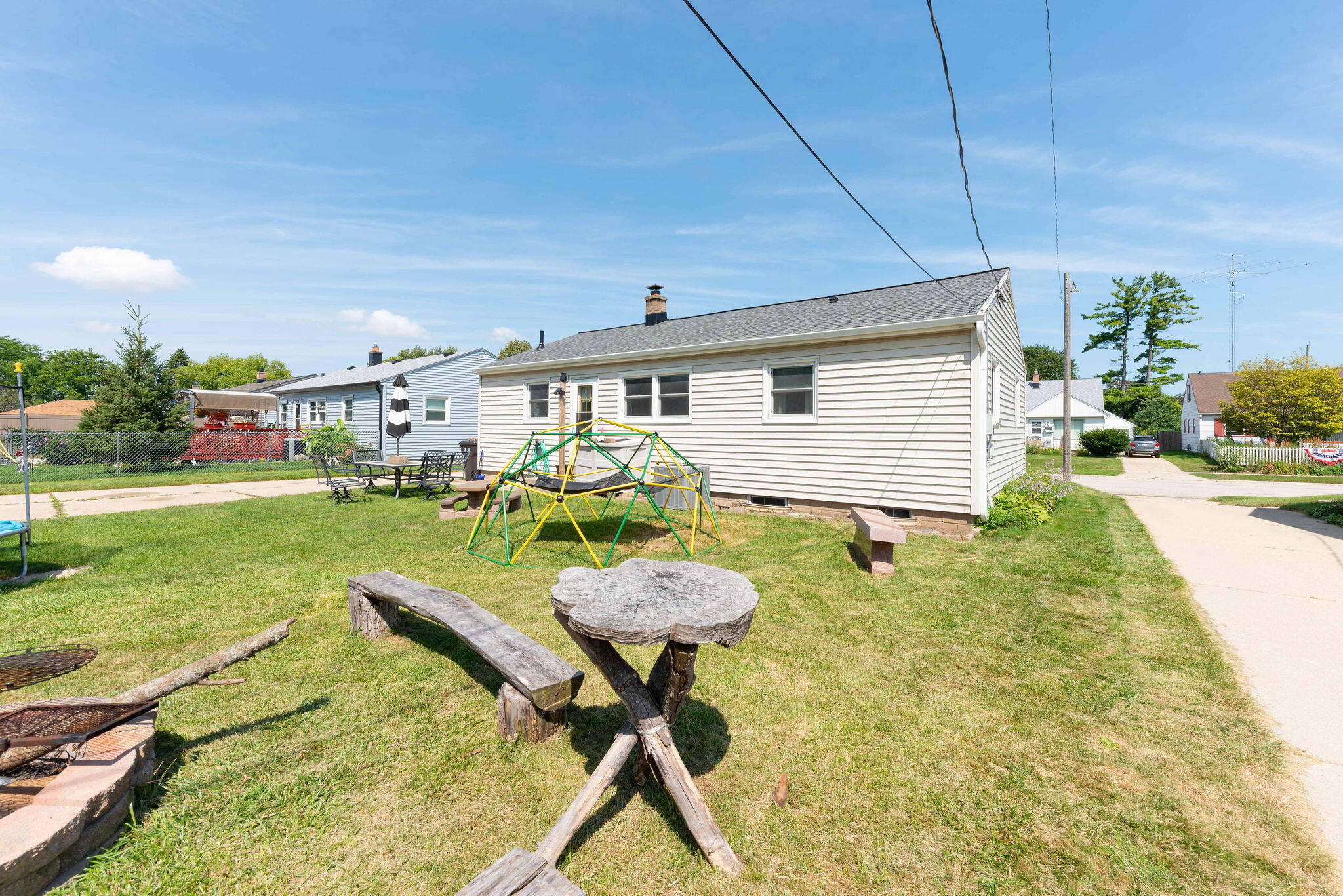 Rear of a white-sided home beyond a backyard with a climbing dome and rustic log seating.