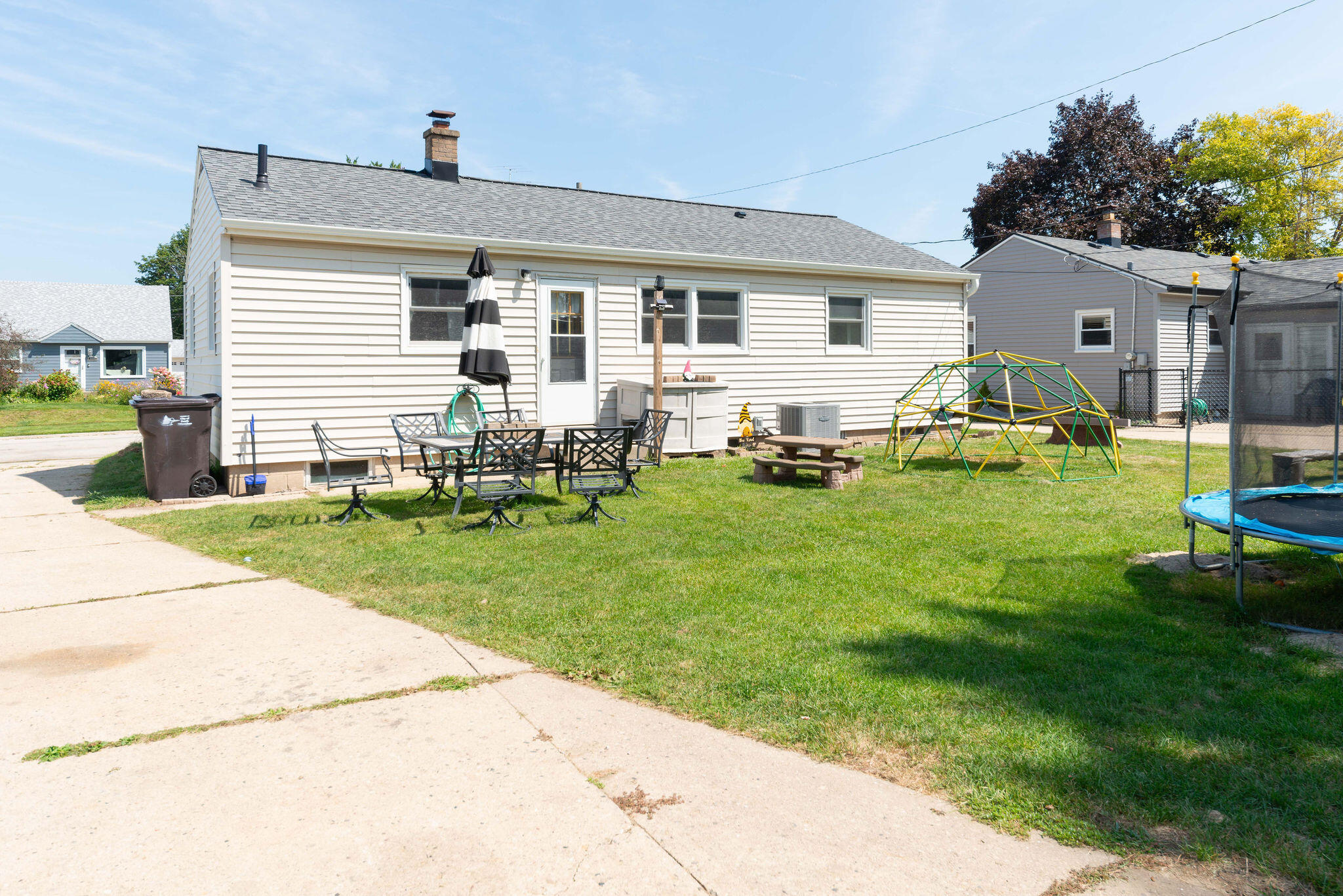 White-sided house and backyard with patio seating, a climbing dome, and trampoline.
