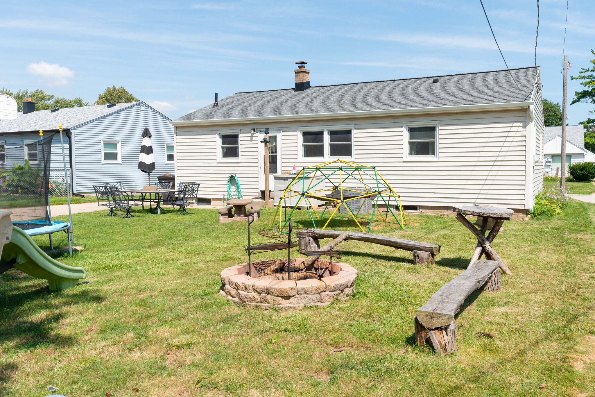 House with a grassy backyard, stone fire pit, play equipment, and outdoor dining set.