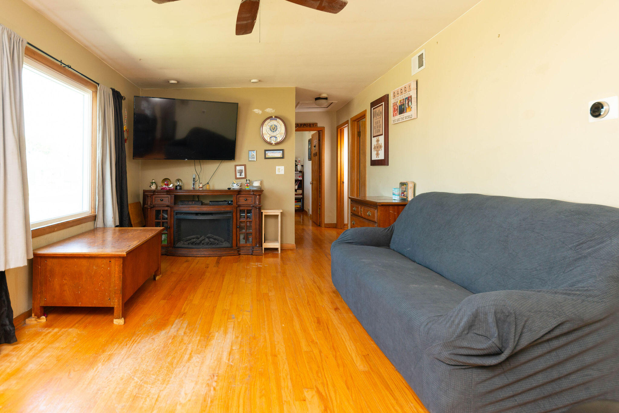 Living room with hardwood floors, a blue-gray sofa, and a wooden fireplace mantel beneath a wall-mounted TV.