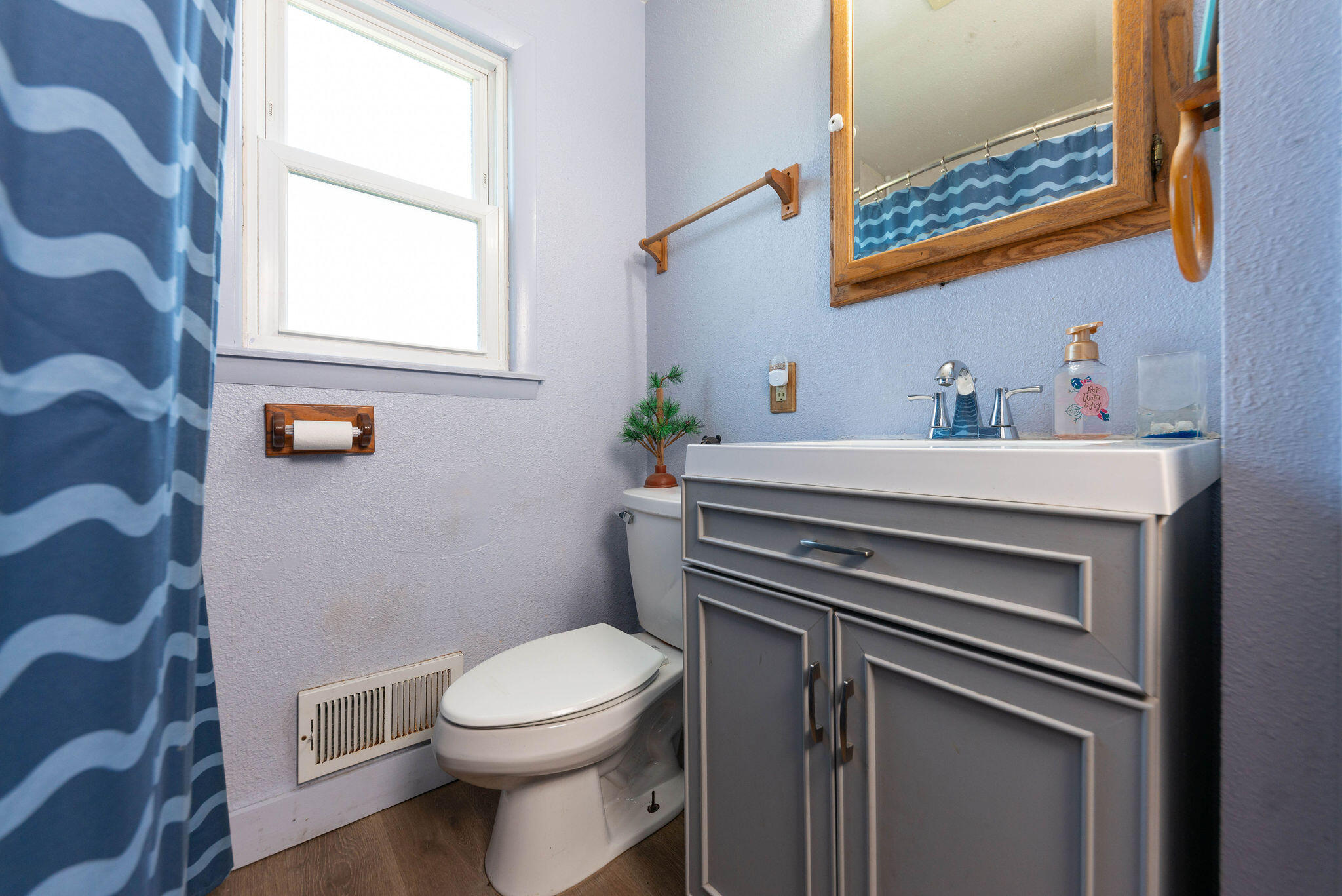 Bathroom with gray vanity and white sink, toilet, blue patterned shower curtain, and bright window.