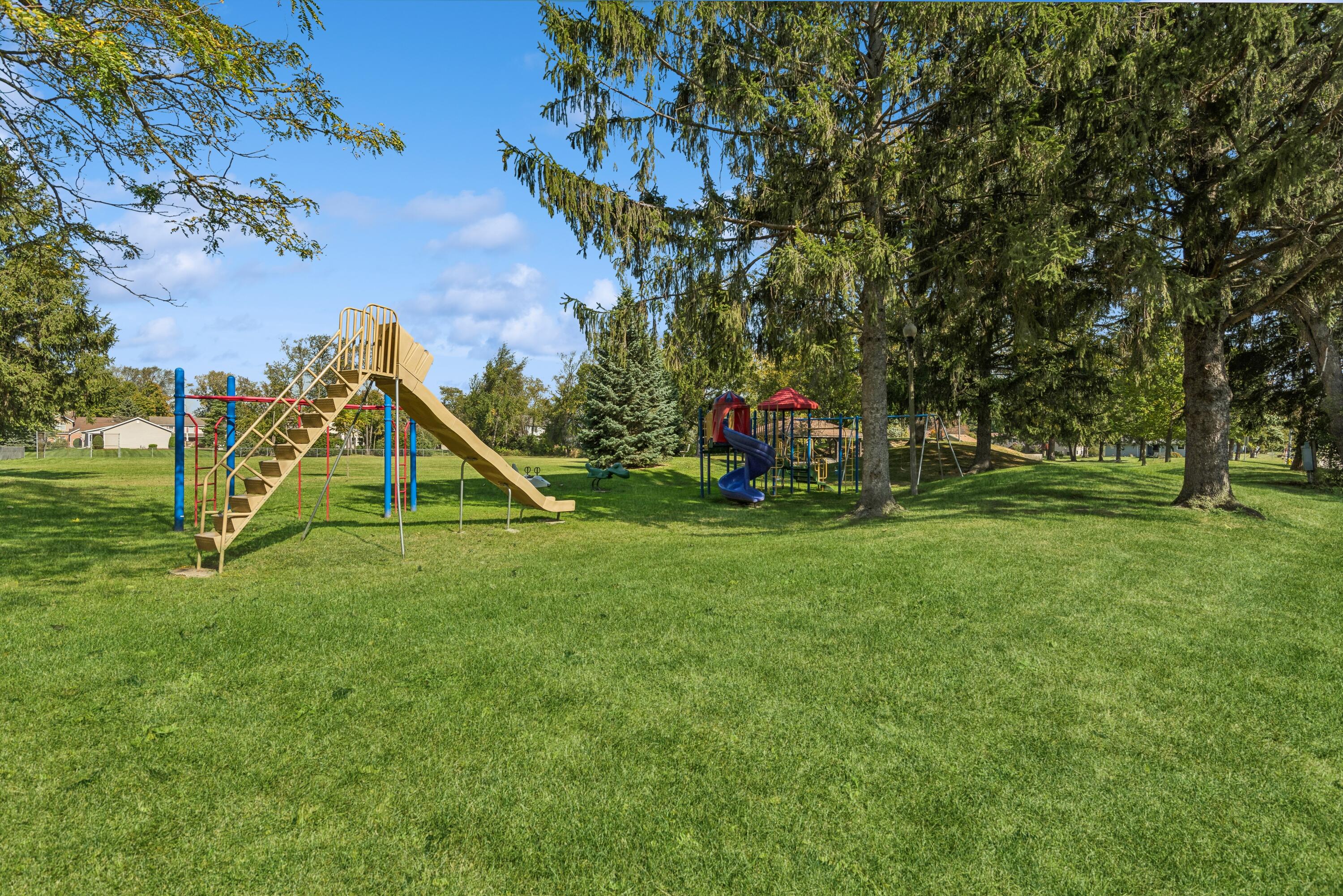 Outdoor playground with two play structures and slides beneath mature trees on a grassy lawn.
