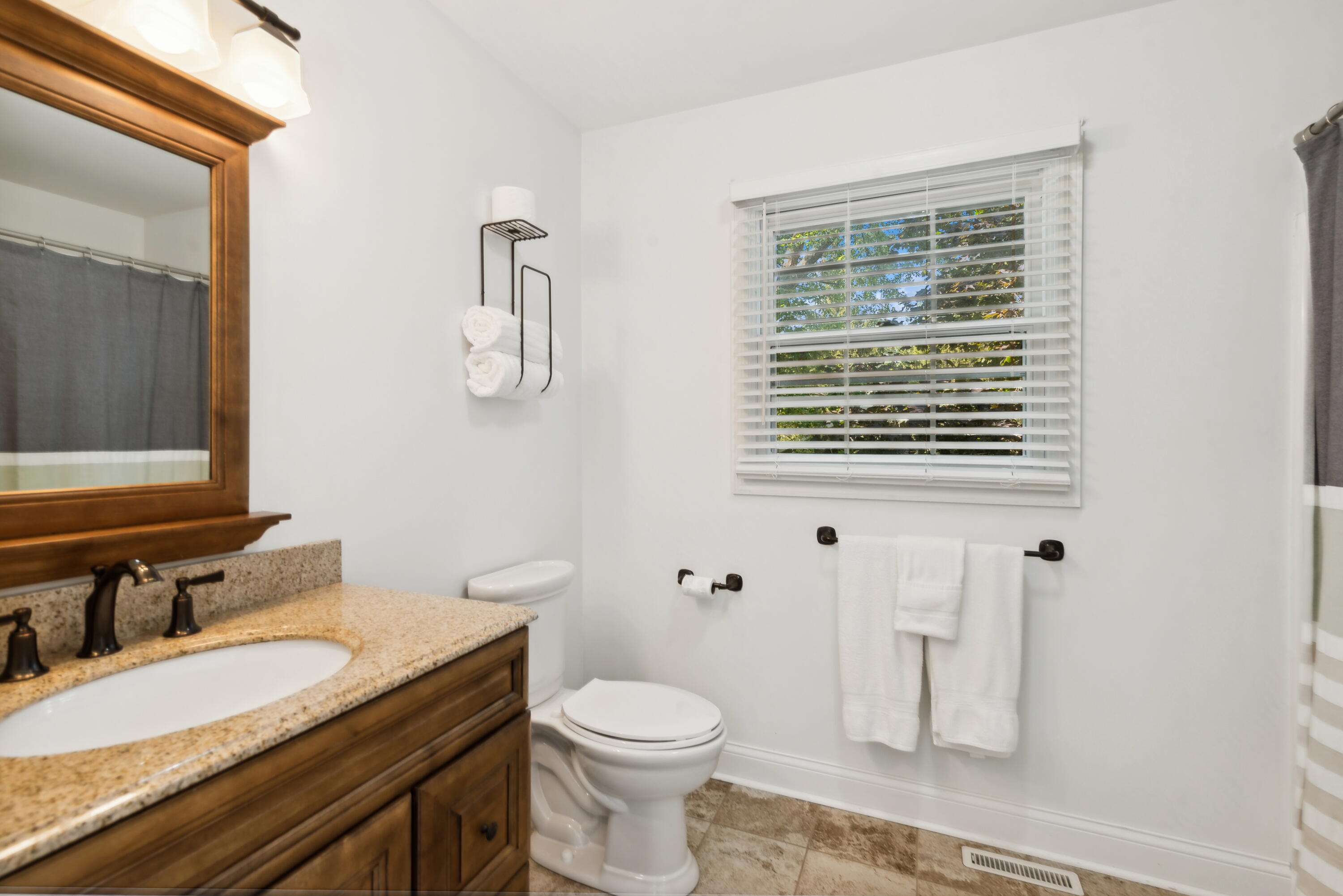 Bathroom with a granite vanity, dark wood cabinetry, white toilet, and window with blinds.