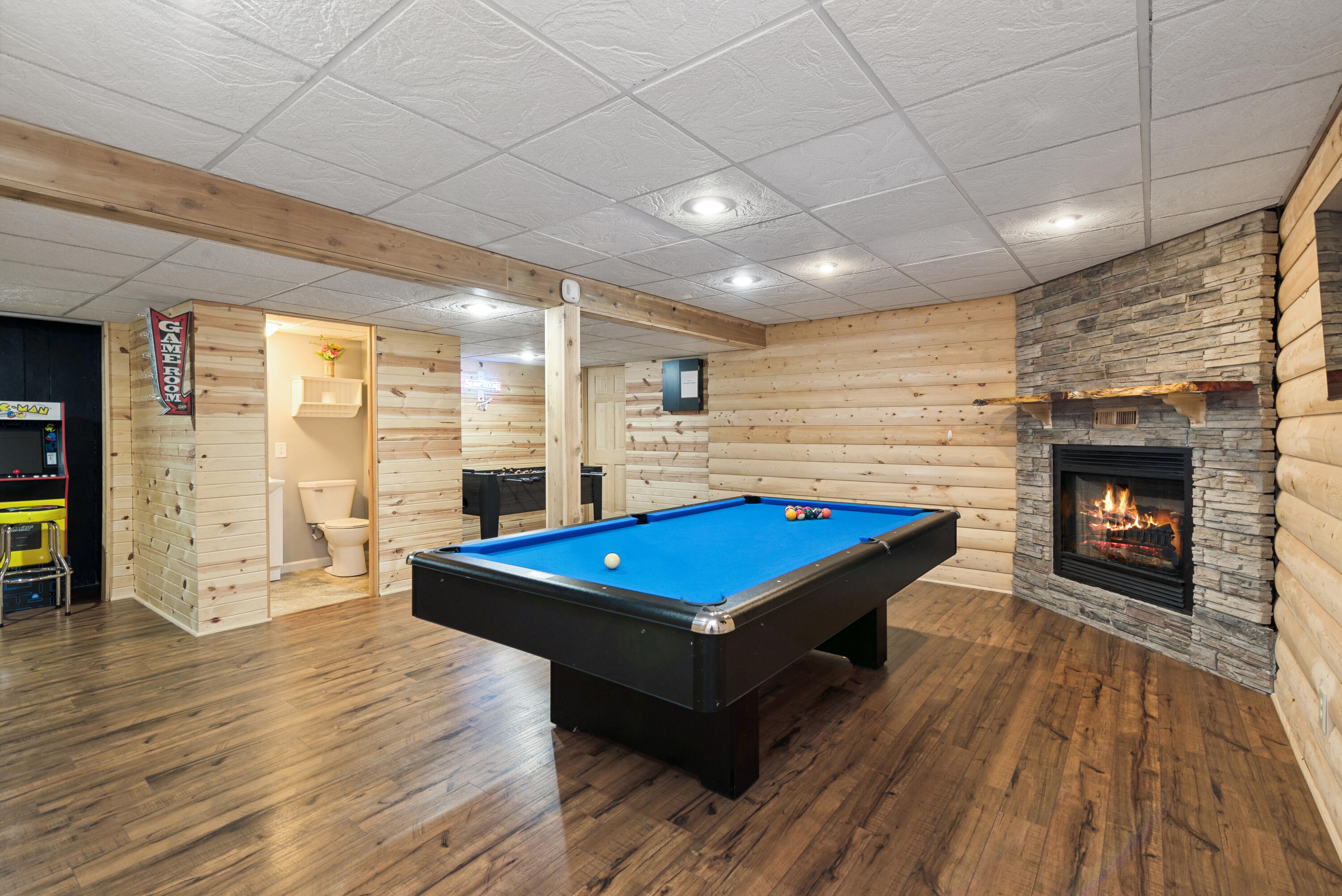 Game room with a blue pool table, stone fireplace, wood-plank walls, and arcade machine.