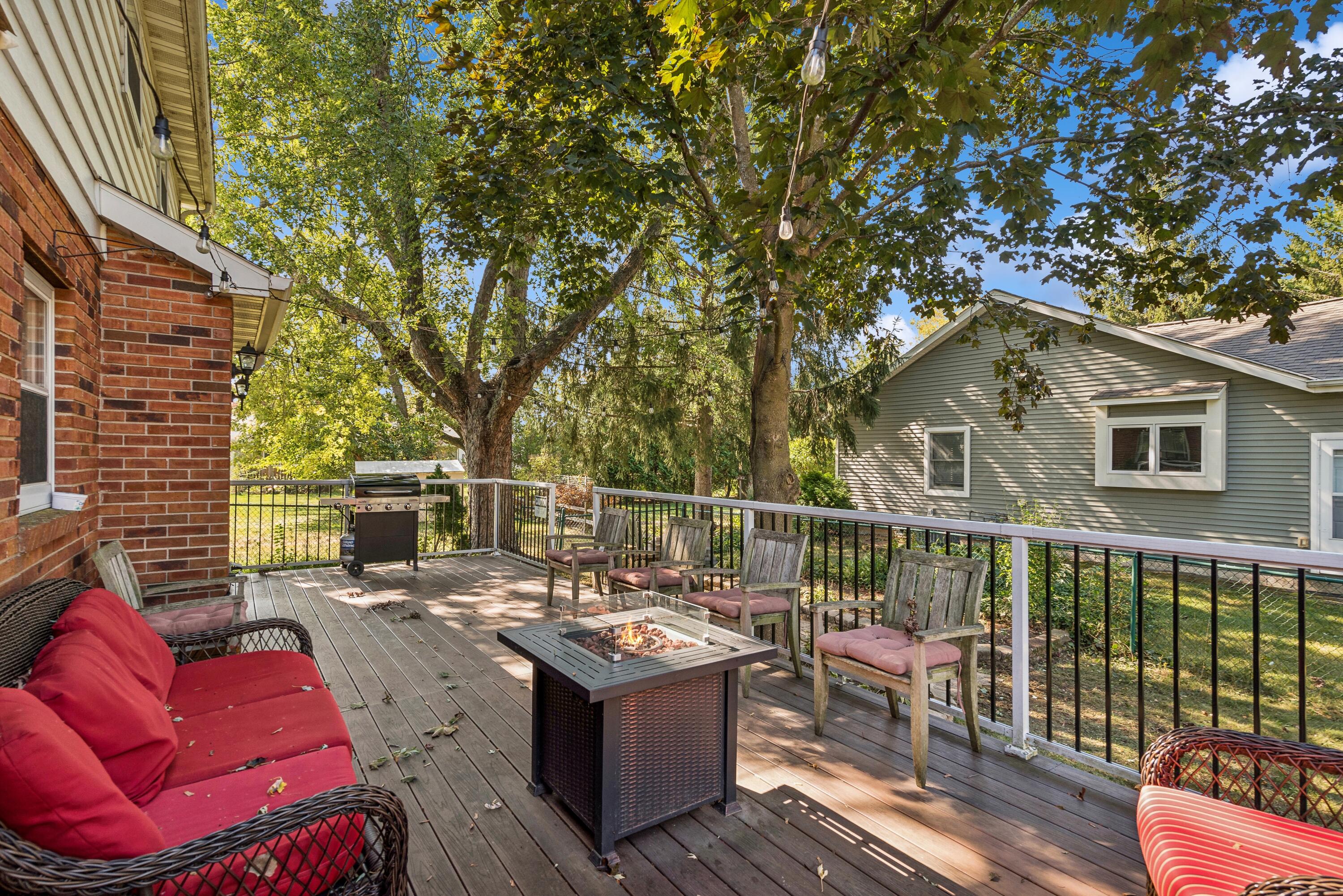 Wooden deck with cushioned seating, a fire table, and grill beneath mature trees.