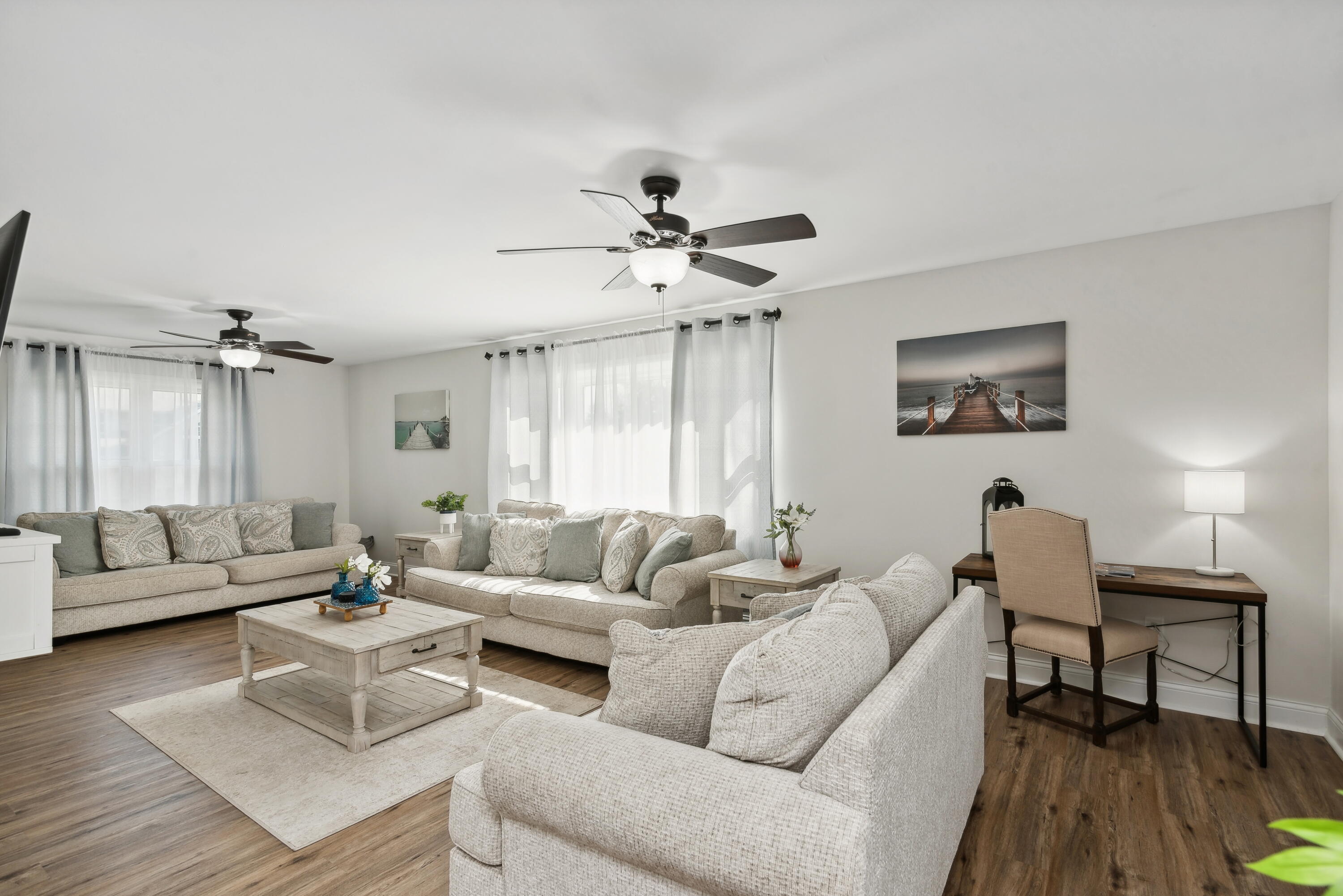 Furnished living room with light sofas, large curtained windows, two ceiling fans, and a desk.