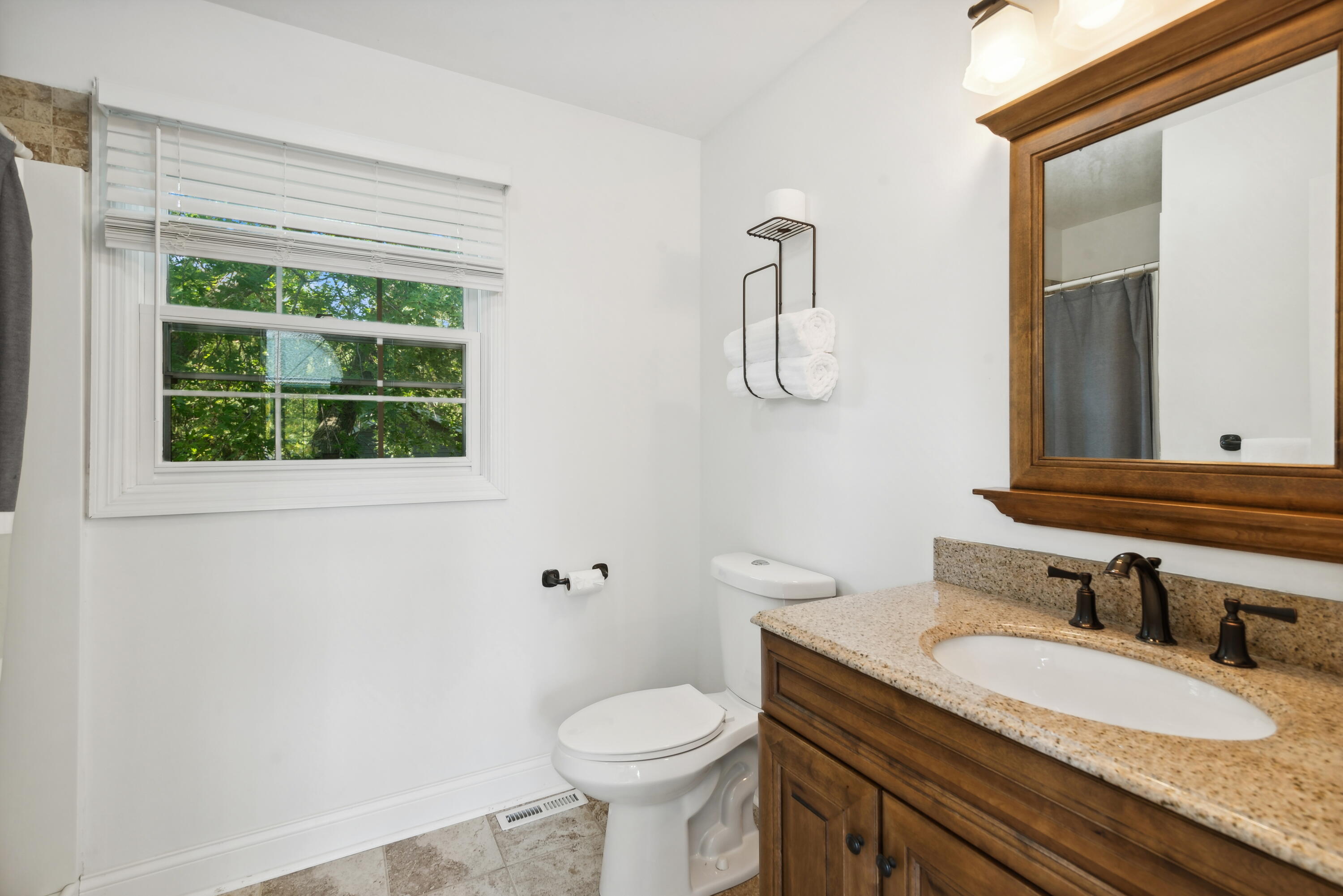 Bathroom with a wood vanity and granite counter, toilet beside a window with leafy trees outside.