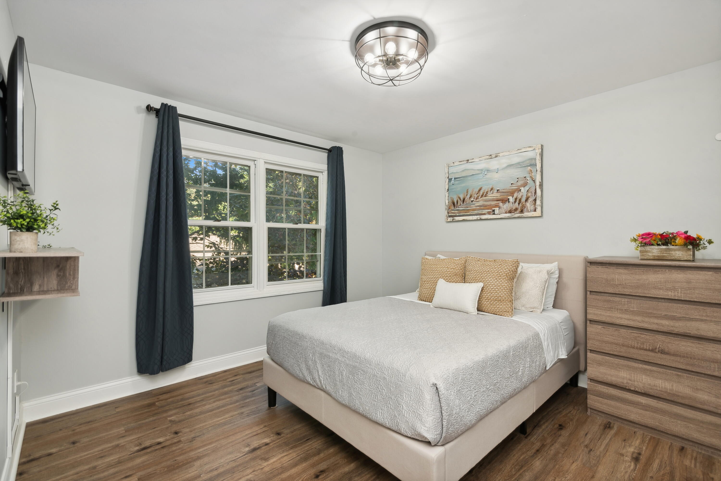 Staged bedroom with an upholstered bed, paired windows with dark curtains, and wood-look flooring.