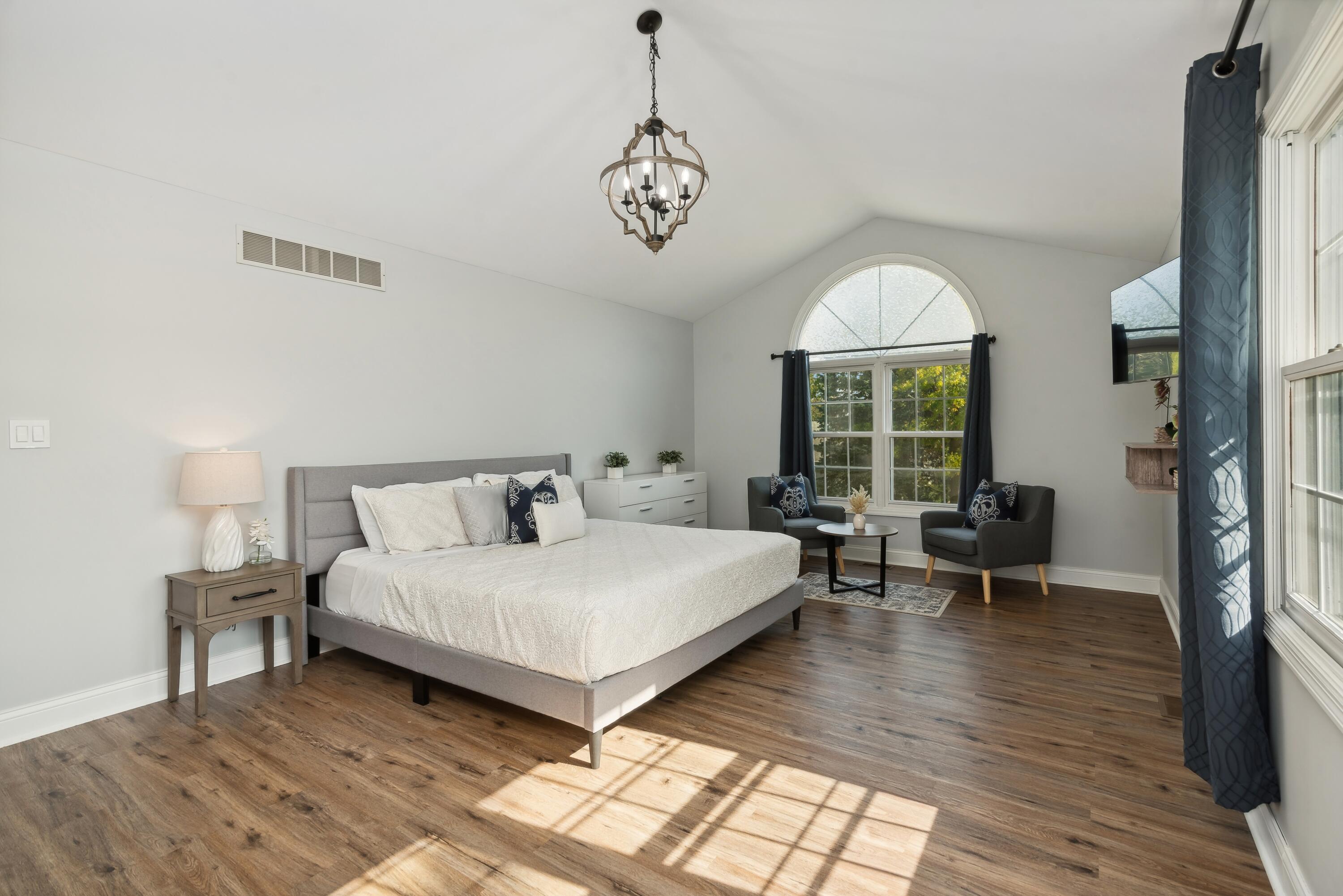 Staged bedroom with a gray bed, arched window, sitting area, and sunlit wood floors.