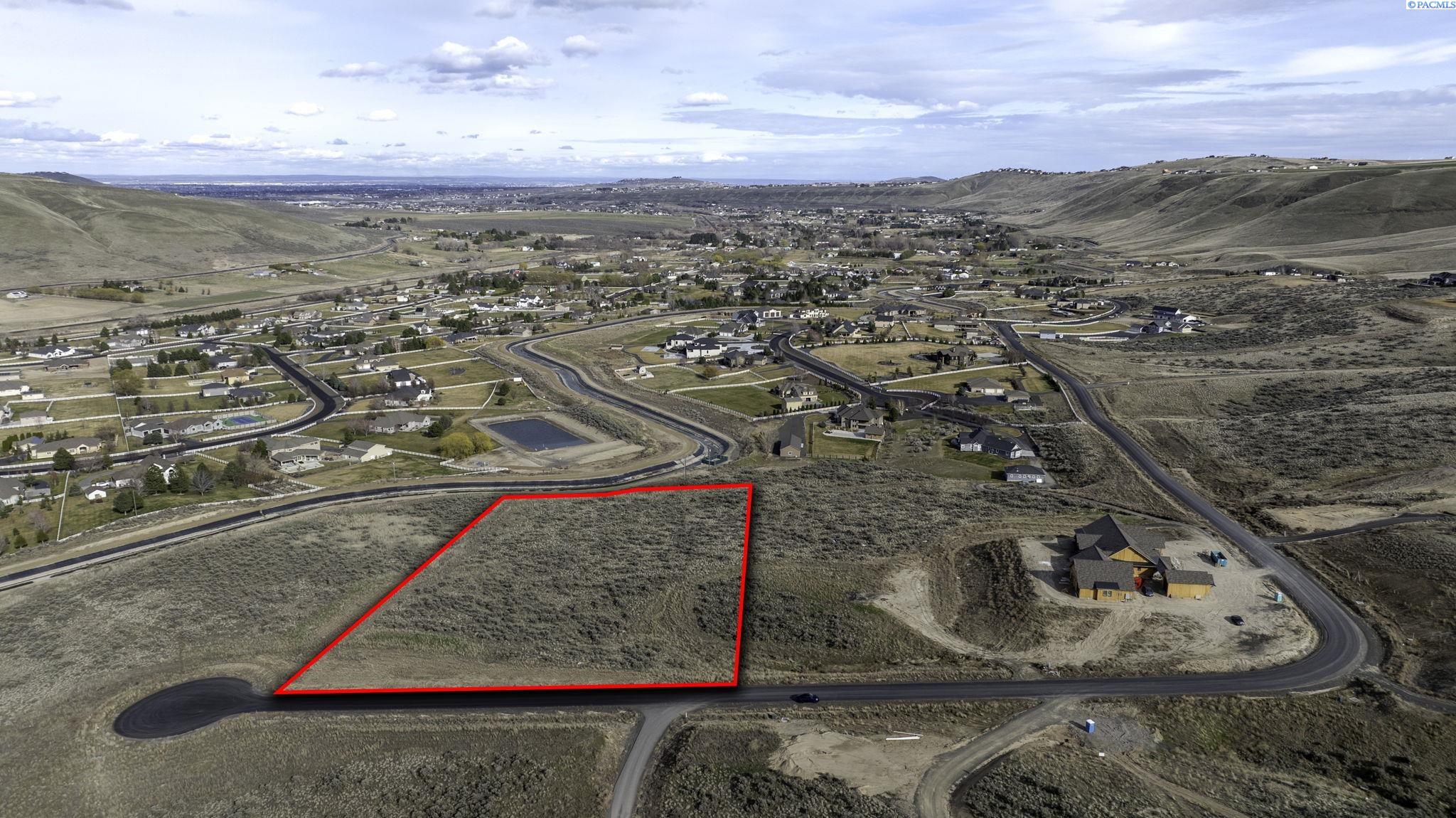 Aerial view of a vacant, red-outlined parcel beside a curved road in a developing hillside neighborhood.