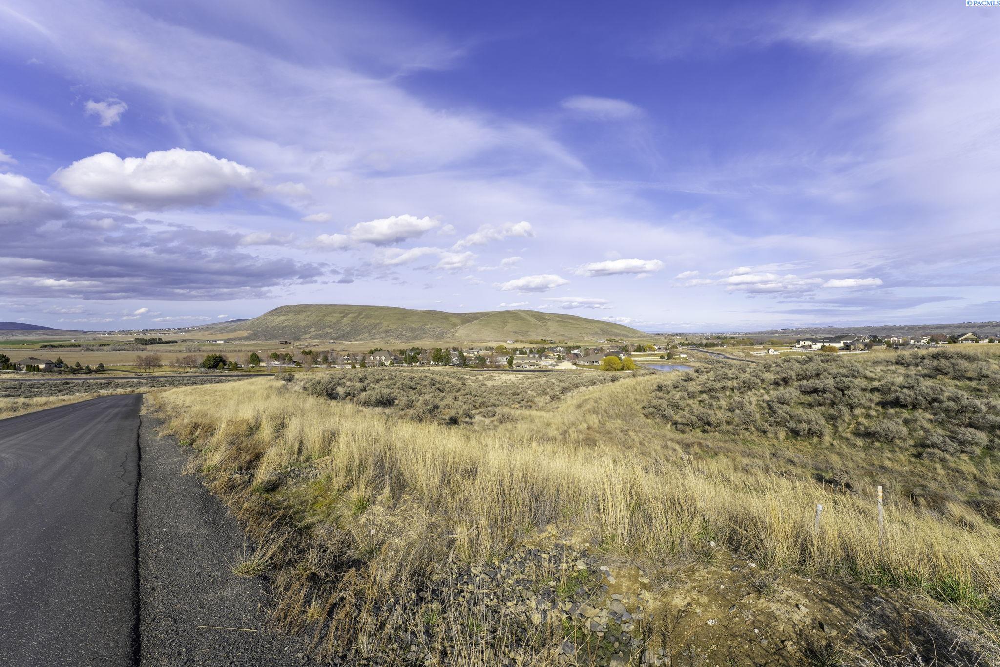Vacant grassy property beside a paved road, overlooking rolling hills and distant homes.