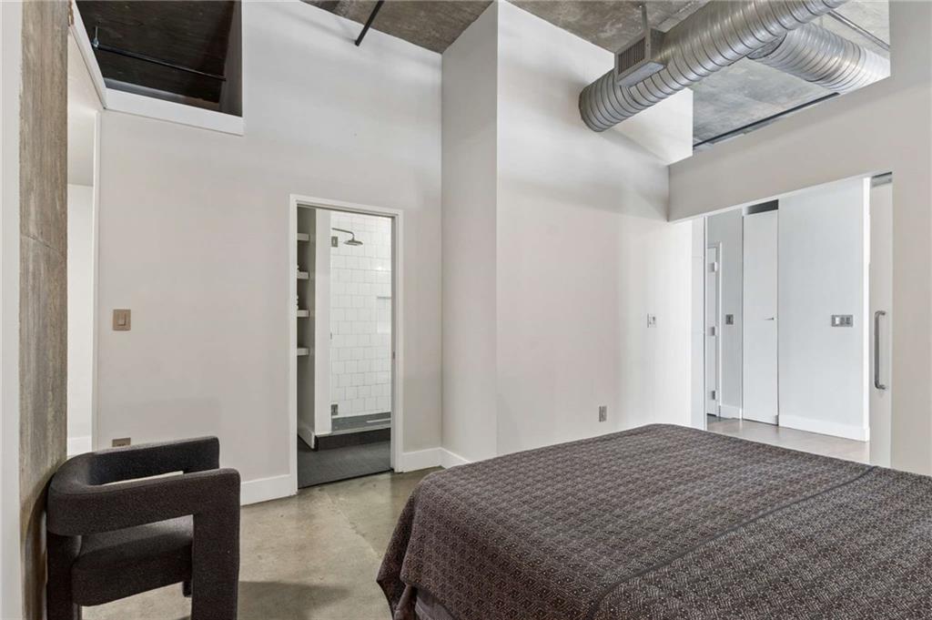 Bedroom with a patterned bedspread, polished concrete floor, exposed ductwork, and a view into the tiled shower.