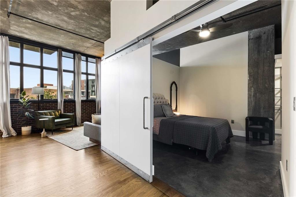 Loft living area and bedroom divided by a white sliding barn door, with tall windows and exposed concrete ceilings.