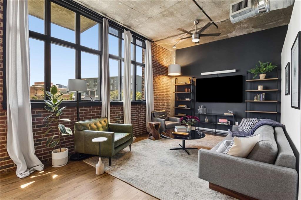 Loft living room with tall windows, exposed brick, and a seating area facing a wall-mounted television.