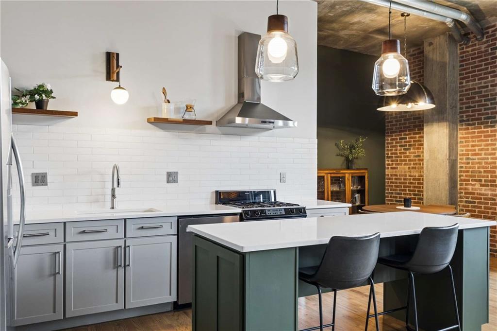 Loft kitchen with a green island, gray cabinetry, white tile backsplash, and exposed brick nearby.