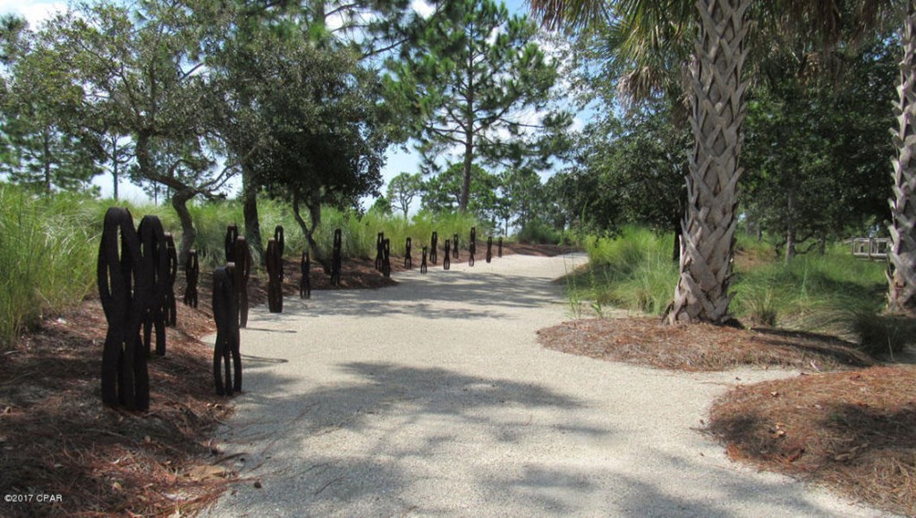 Gravel path lined with dark metal figure sculptures beneath trees and palms.