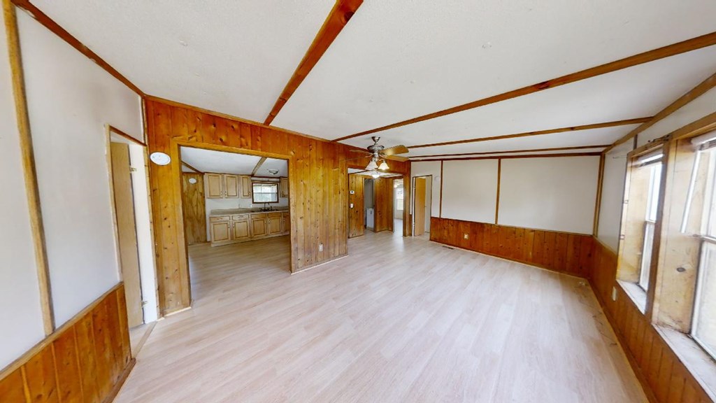 Empty living room with wood paneling, pale floors, and an open view into the kitchen.