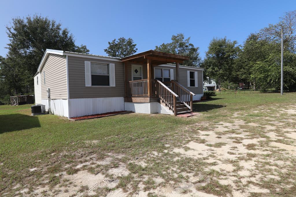 Manufactured home with a covered wooden porch and stairs in a grassy, sandy yard.