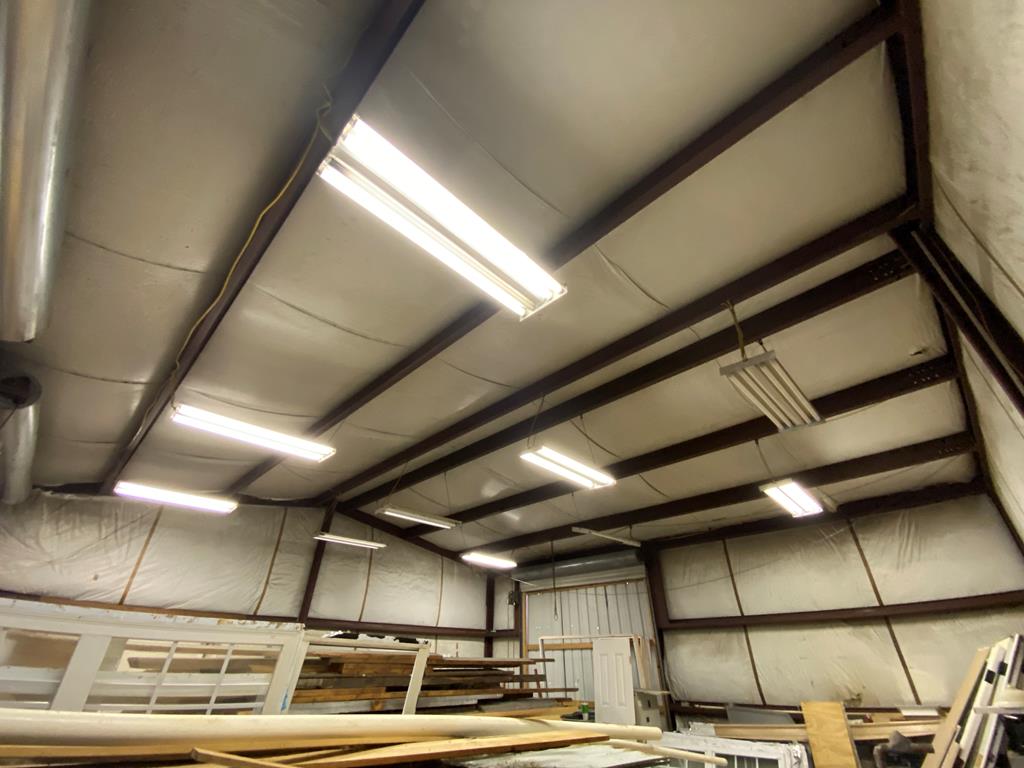 Workshop interior with exposed steel trusses, fluorescent lights, and lumber storage.