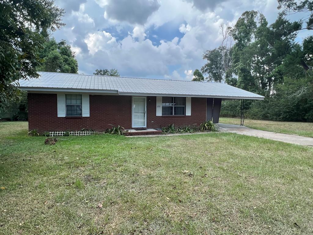 Single-story brick house with a light metal roof, attached carport, and broad grassy lawn.